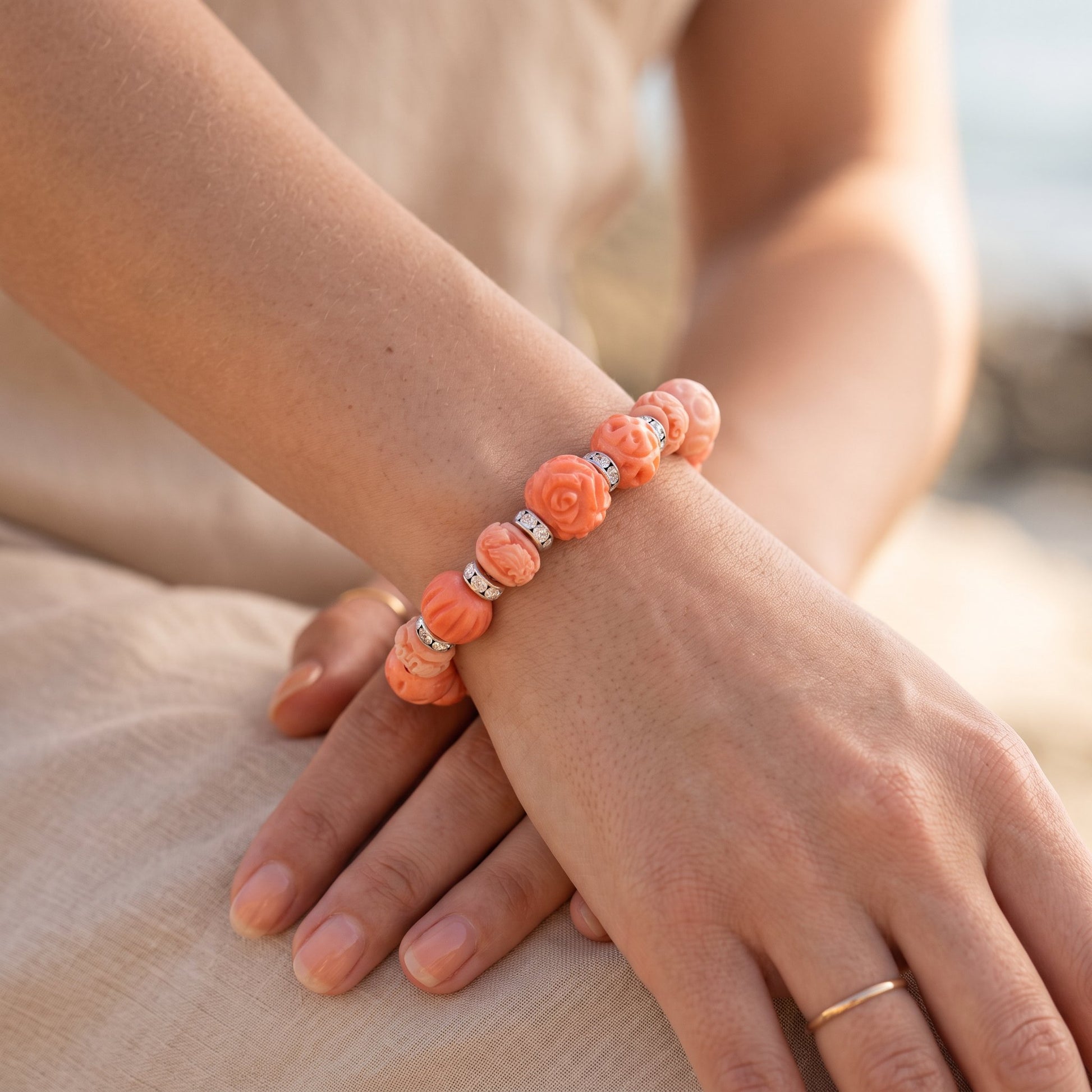A close-up of a person's hands and wrist wearing a coral-orange beaded bracelet with rose-shaped beads and small silver spacer accents, set against a soft beige background.