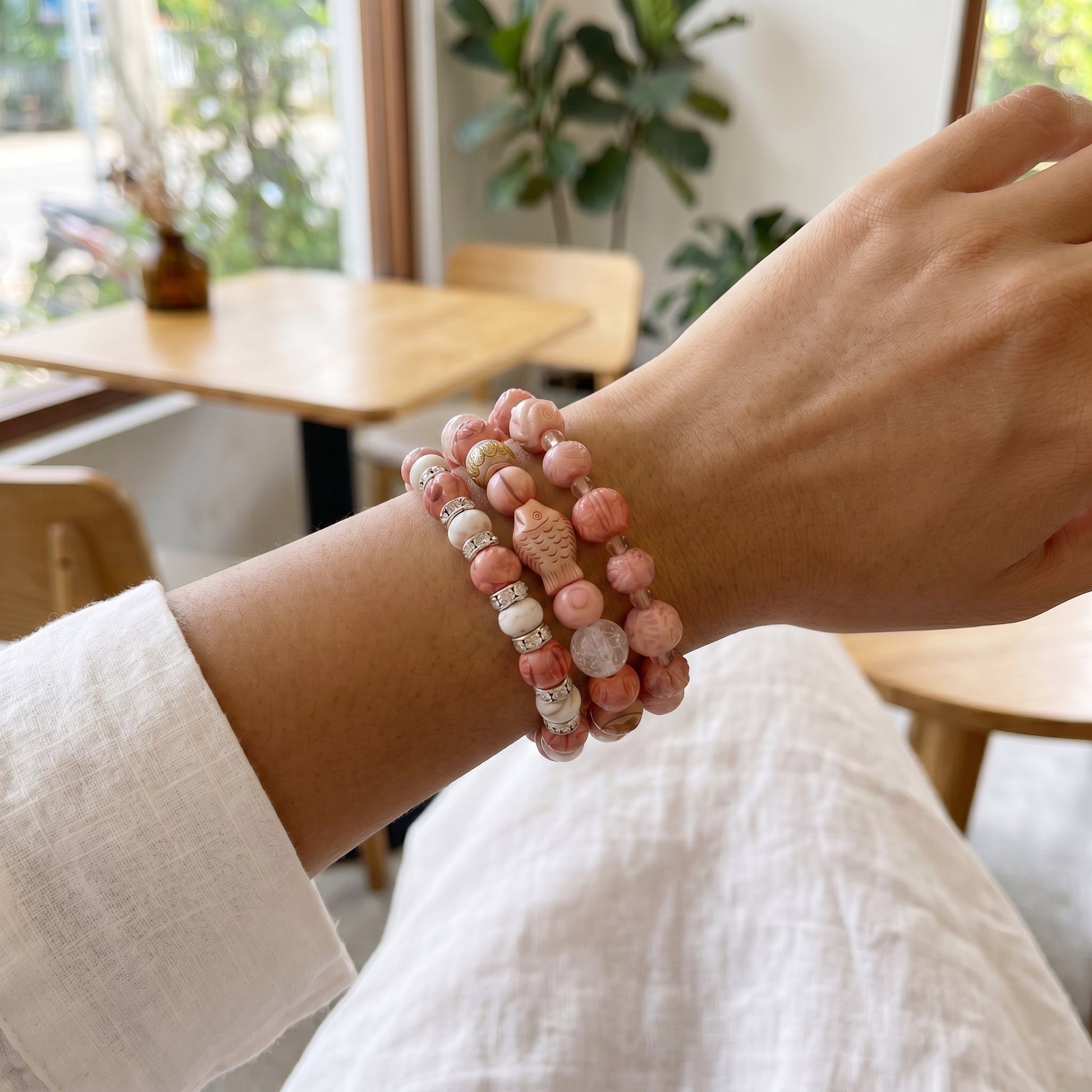 A close-up of a person's wrist wearing three beaded bracelets in a softly lit indoor setting with wooden furniture and plants in the background.