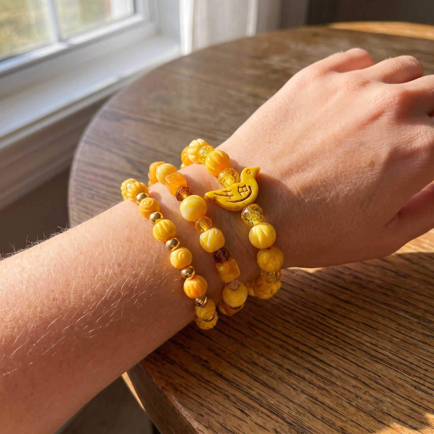 Three yellow beaded stretch bracelets with a small yellow duck charm, worn on a person's wrist resting on a wooden table near a window.