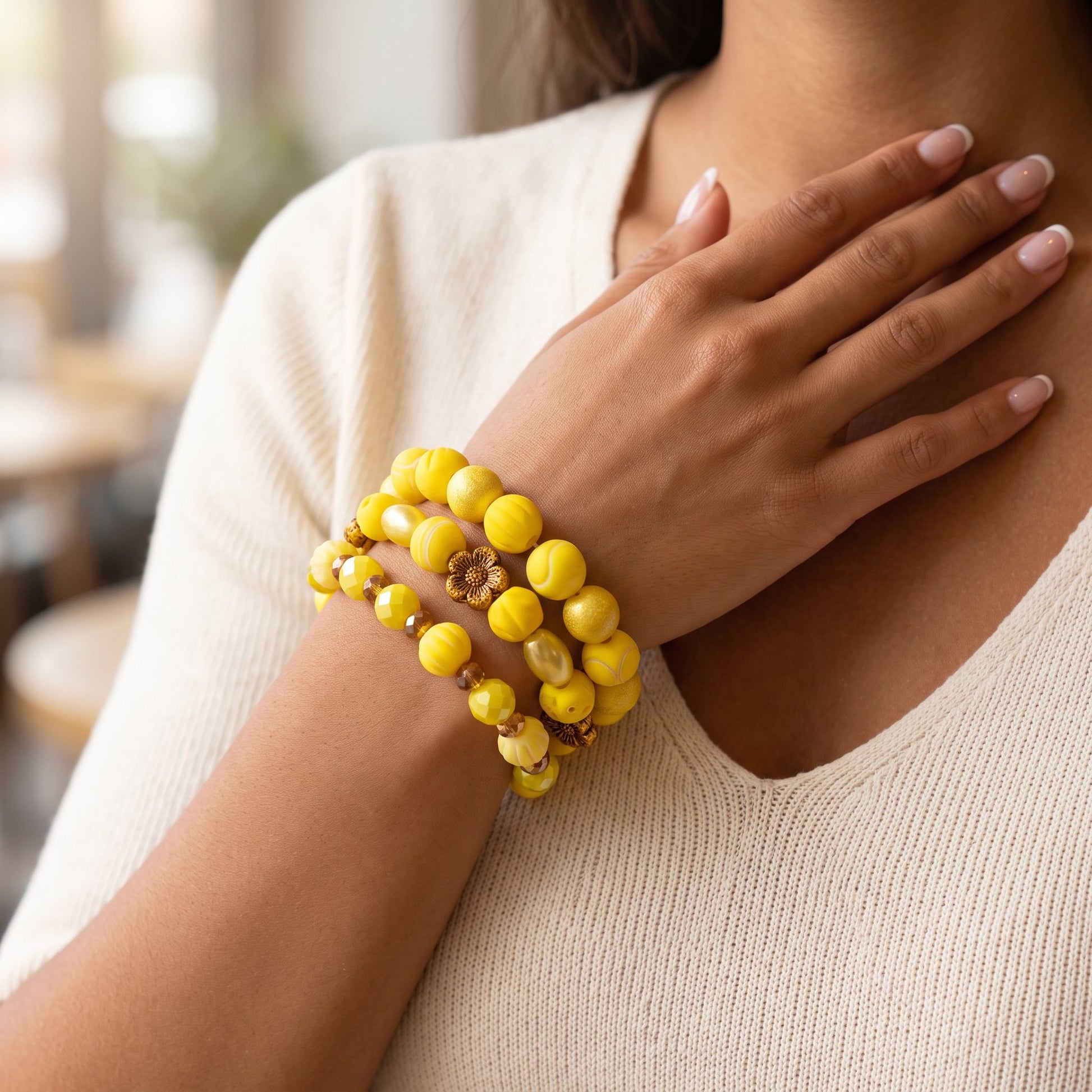 A close-up of a person wearing three yellow beaded bracelets on their wrist, including one with a small brown floral charm, against a soft beige knit top.