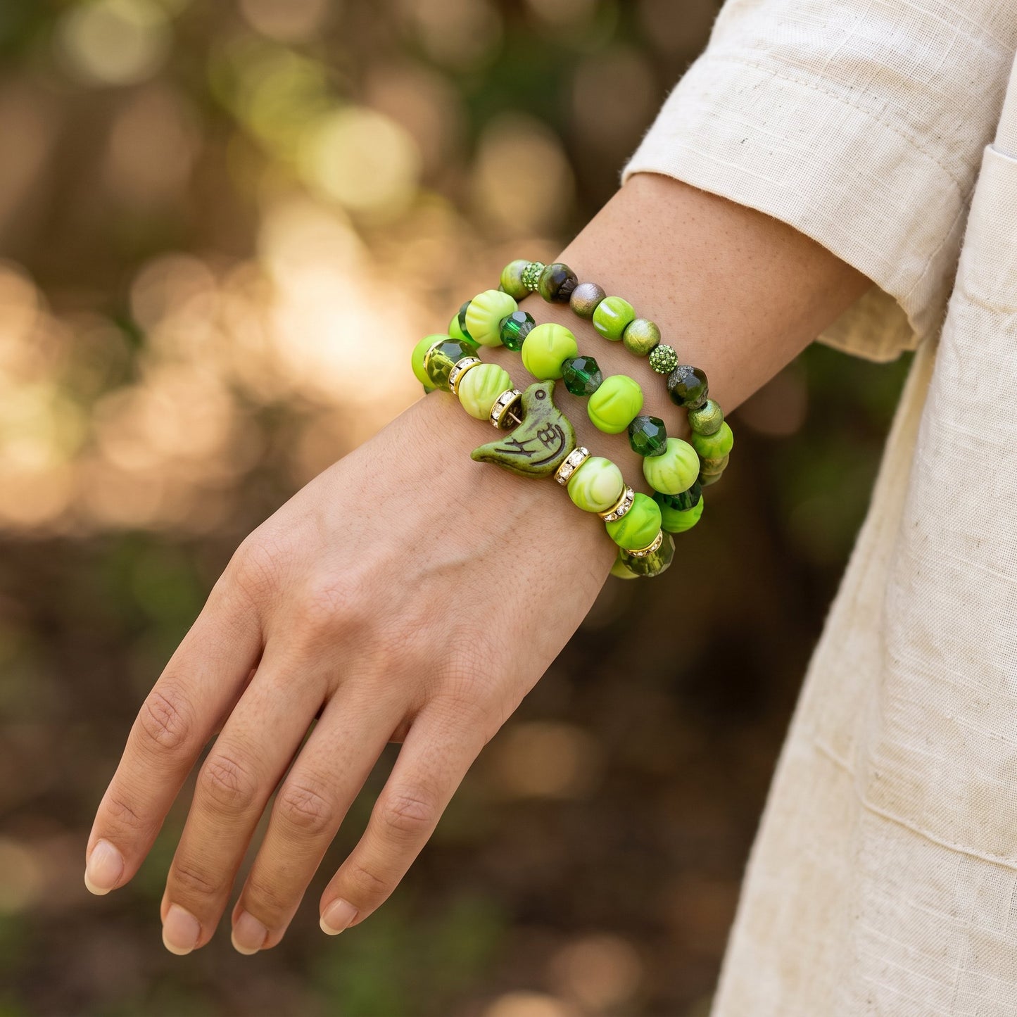 A close-up of a person's wrist wearing three stacked beaded bracelets. The bracelets feature light green and dark green marbled beads, with one bracelet decorated with a small green leaf charm and gold spacer beads.