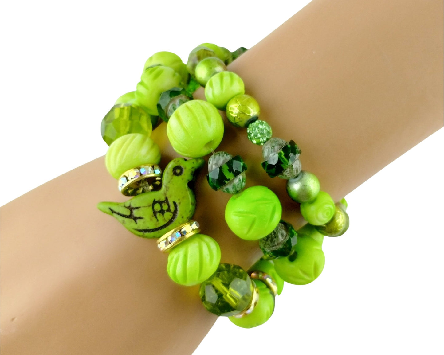 A close-up of a person's wrist wearing a stack of three beaded bracelets. The bracelets feature bright lime green and emerald green beads, faceted green crystal beads, and a green bird charm with black details and small gold rhinestone accents.