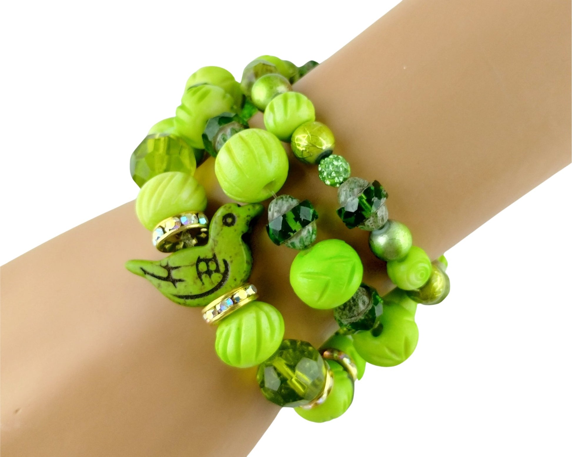 A close-up of a person's wrist wearing a stack of three beaded bracelets. The bracelets feature bright lime green and emerald green beads, faceted green crystal beads, and a green bird charm with black details and small gold rhinestone accents.