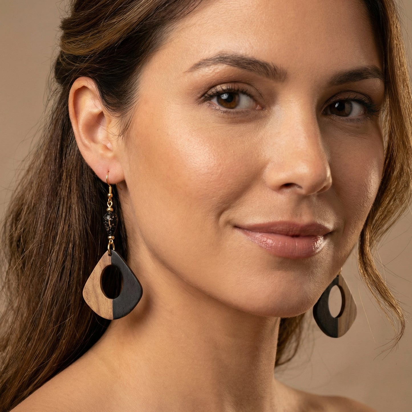 Close-up of a woman wearing black and wood  earrings with gold findings against a neutral background