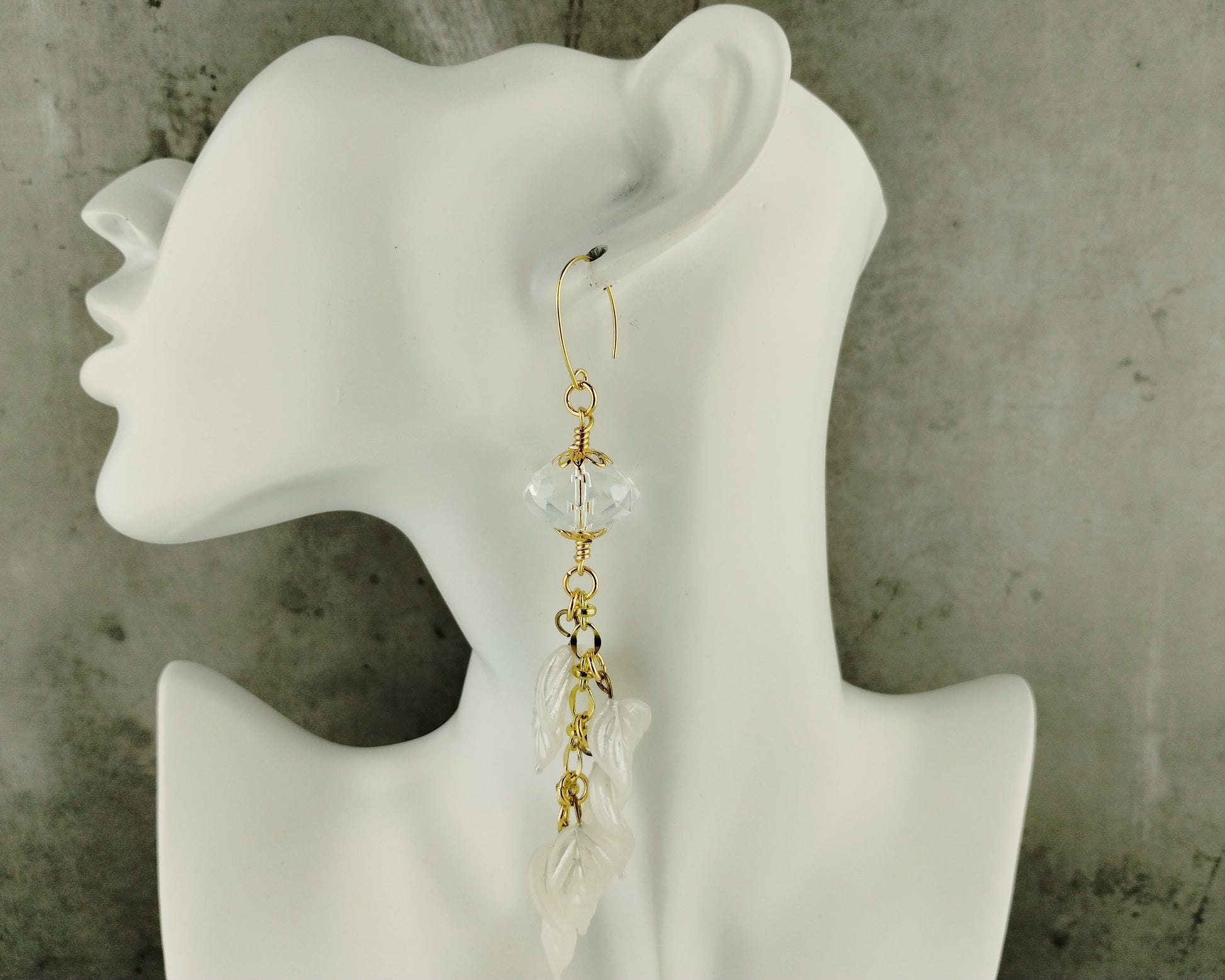 Gold and white dangling earrings on a white mannequin ear against a gray background
