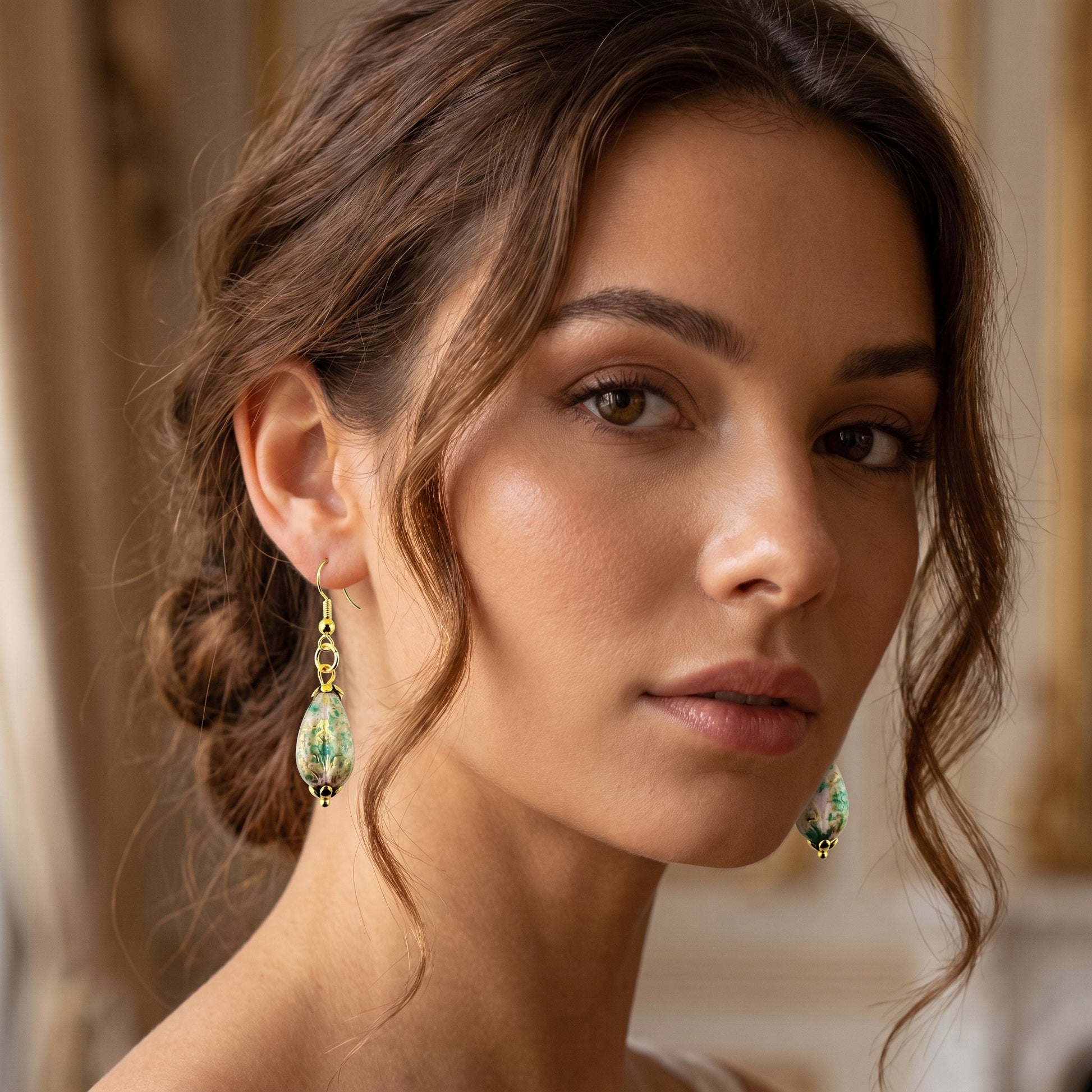A close-up of a woman wearing gold teardrop dangle earrings with green and white marbled stone-like drops, set against a softly blurred warm beige interior background.