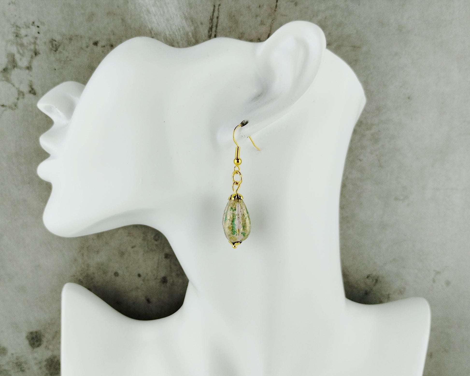A gold dangle earring with a teardrop-shaped clear resin pendant containing green and gold leaf details, displayed on a white mannequin head against a textured gray concrete background.