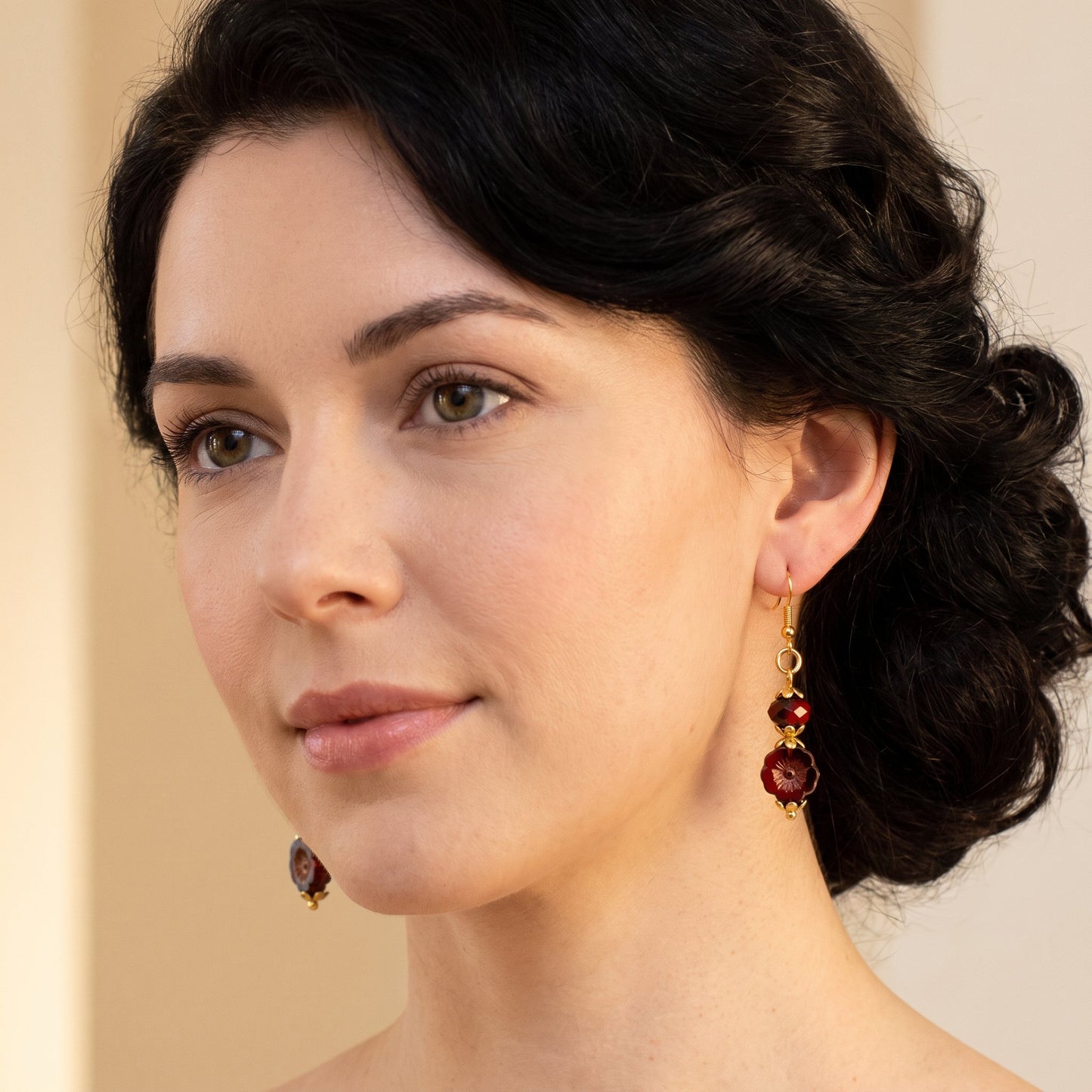 A close-up portrait of a woman wearing dangling earrings with faceted dark red gemstones and gold-toned metal details.