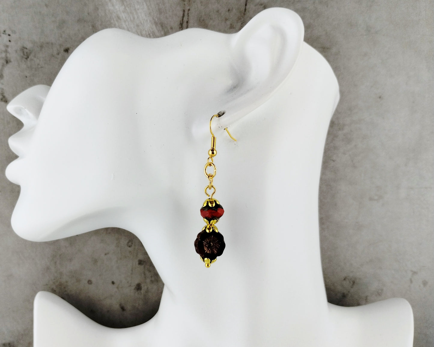 A single dangling earring displayed on a white female mannequin head against a gray concrete-textured background. The earring has a gold-tone fishhook and chain, with a faceted dark red bead and a larger dark brown or black beaded drop.