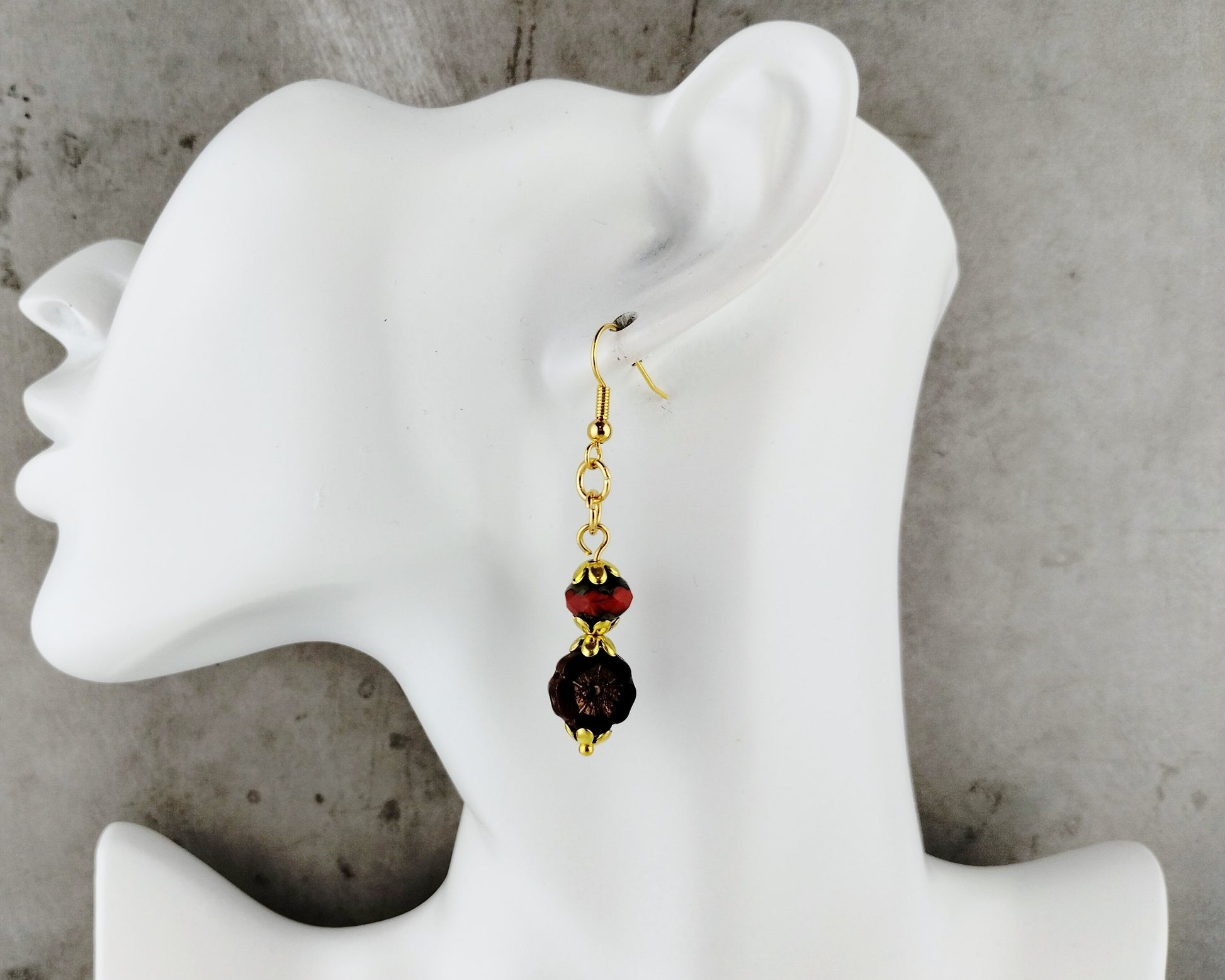 A single dangling earring displayed on a white female mannequin head against a gray concrete-textured background. The earring has a gold-tone fishhook and chain, with a faceted dark red bead and a larger dark brown or black beaded drop.