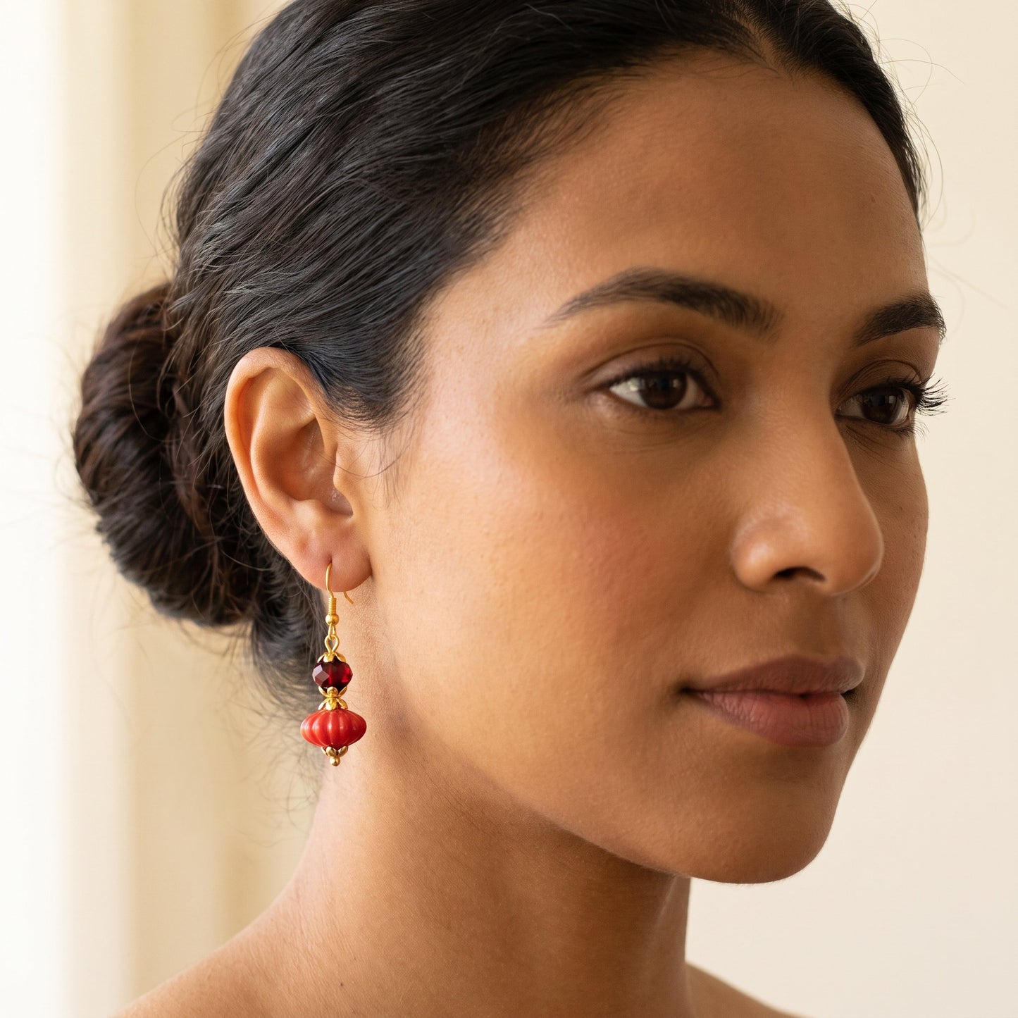 A close-up of a woman wearing a dangling earring with red beads and gold-toned metal hooks, shown against a soft cream background.