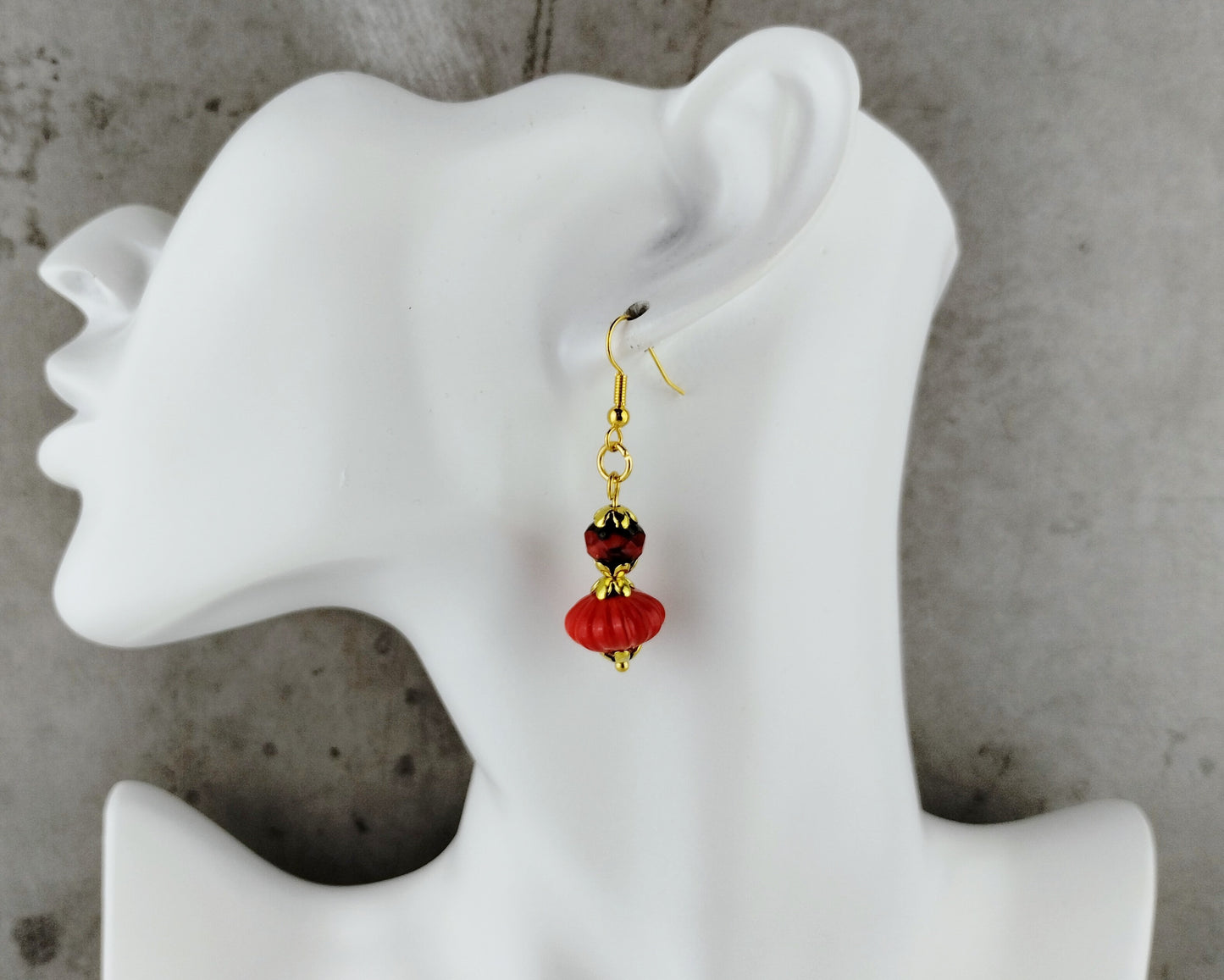 A single dangling earring displayed on a white female mannequin head against a gray concrete-textured background. The earring has gold-tone metal hardware with a dark red faceted bead and a larger red bead below it.