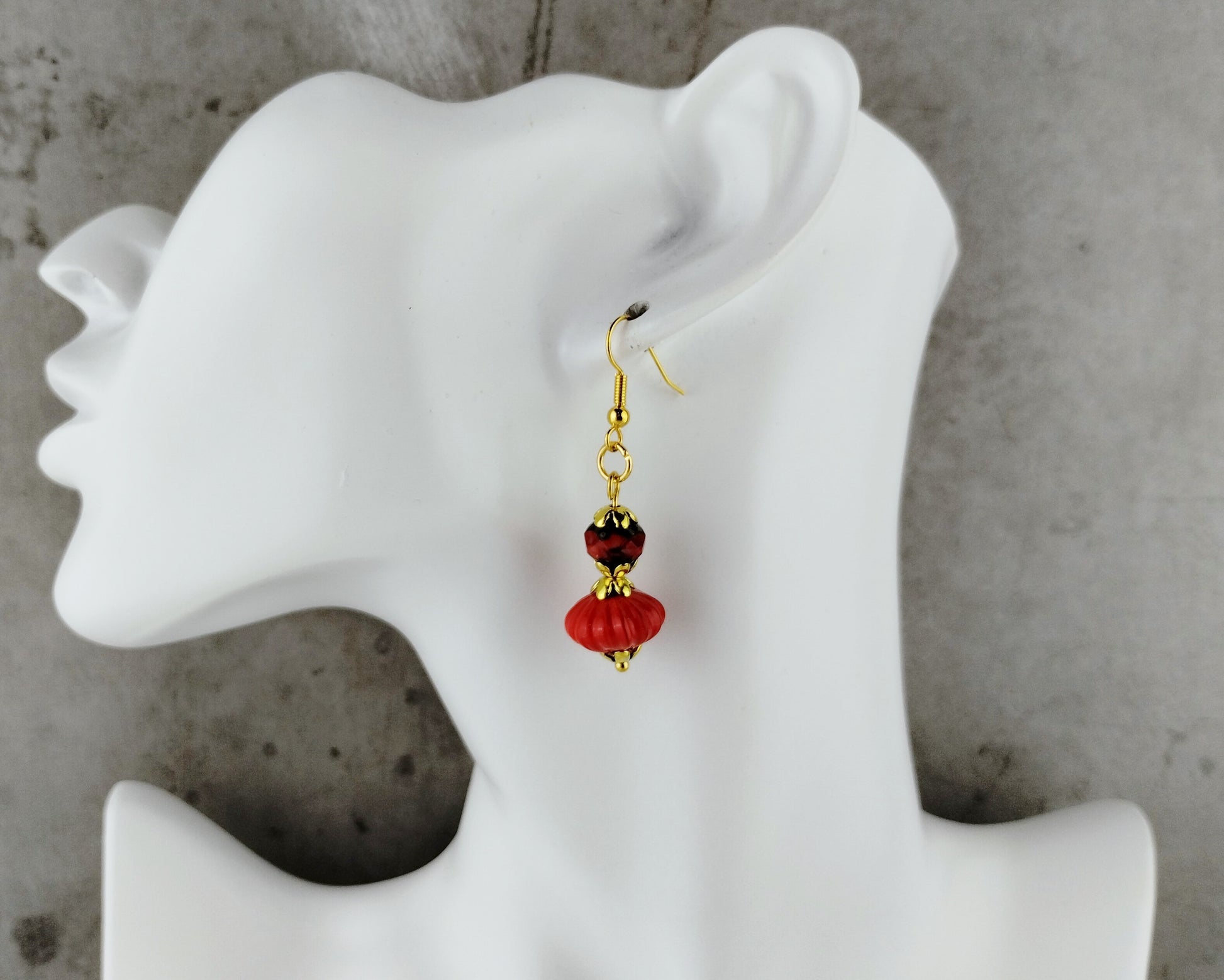 A single dangling earring displayed on a white female mannequin head against a gray concrete-textured background. The earring has gold-tone metal hardware with a dark red faceted bead and a larger red bead below it.