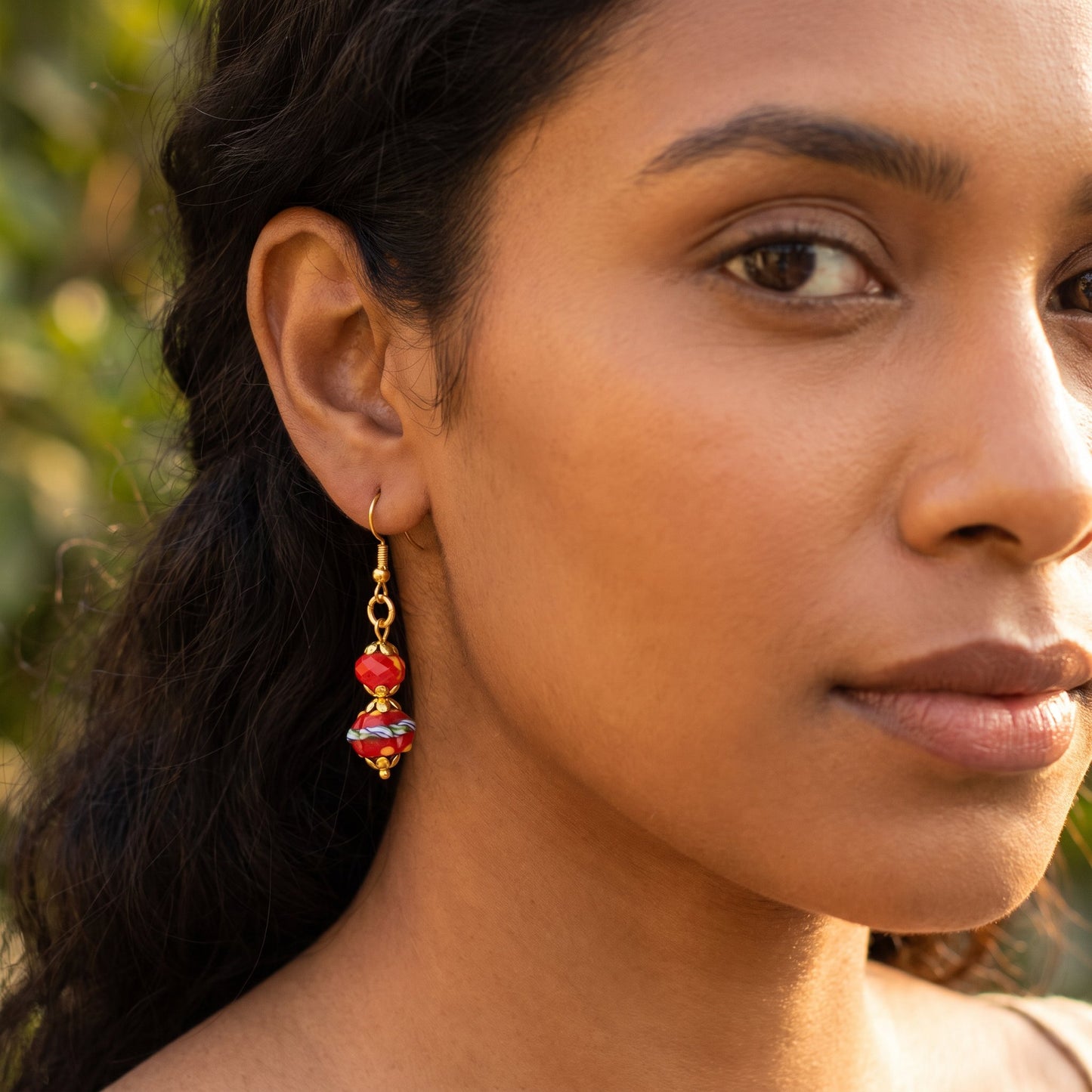 A close-up of a woman wearing a dangling glass earring with red and white marbled beads on a gold-tone hook, photographed outdoors with soft green bokeh in the background.
