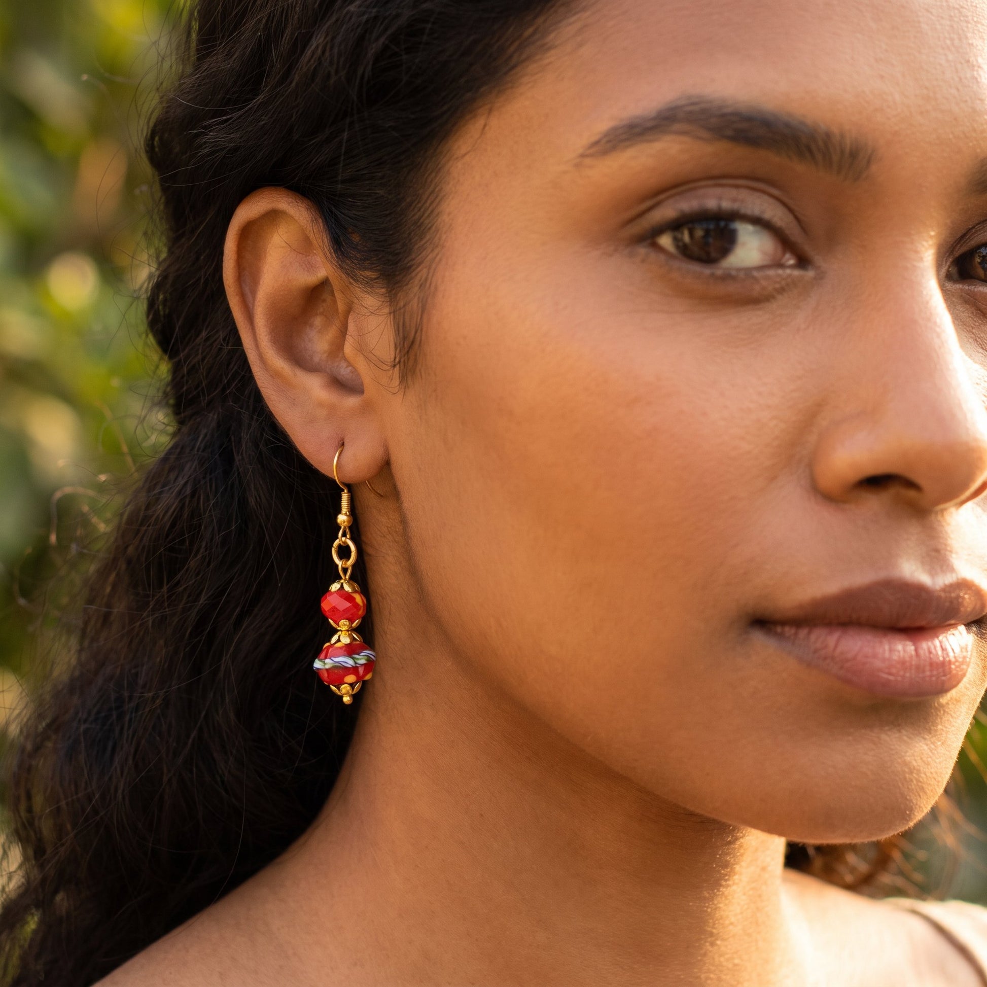A close-up of a woman wearing a dangling glass earring with red and white marbled beads on a gold-tone hook, photographed outdoors with soft green bokeh in the background.