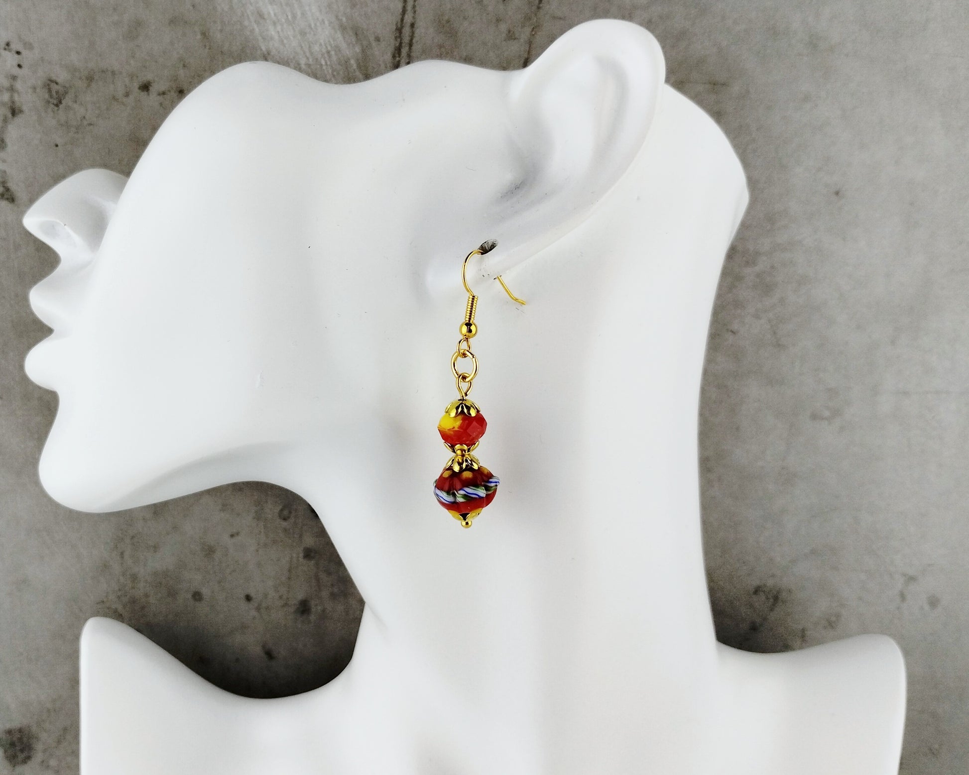 A single dangling earring displayed on a white mannequin head against a gray concrete-textured background. The earring has a gold-tone hook and bead chain with two amber-orange beads and a smaller iridescent purple-blue bead at the bottom.