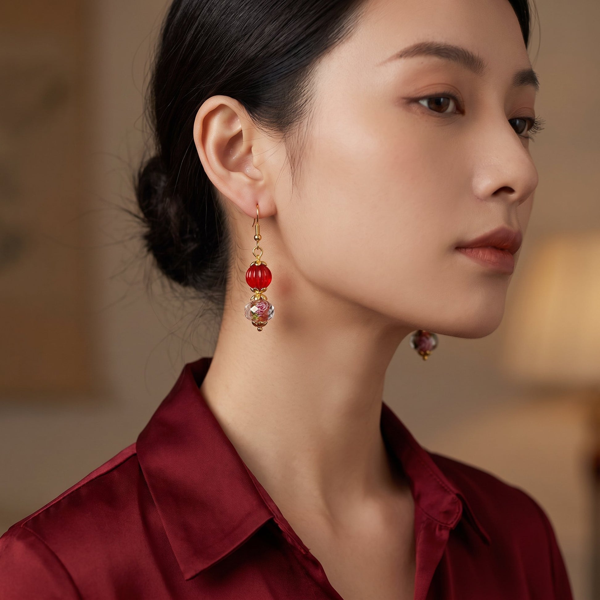 A close-up of a woman wearing a pair of dangling earrings with red beads and gold-toned metal details, paired with a burgundy satin shirt.