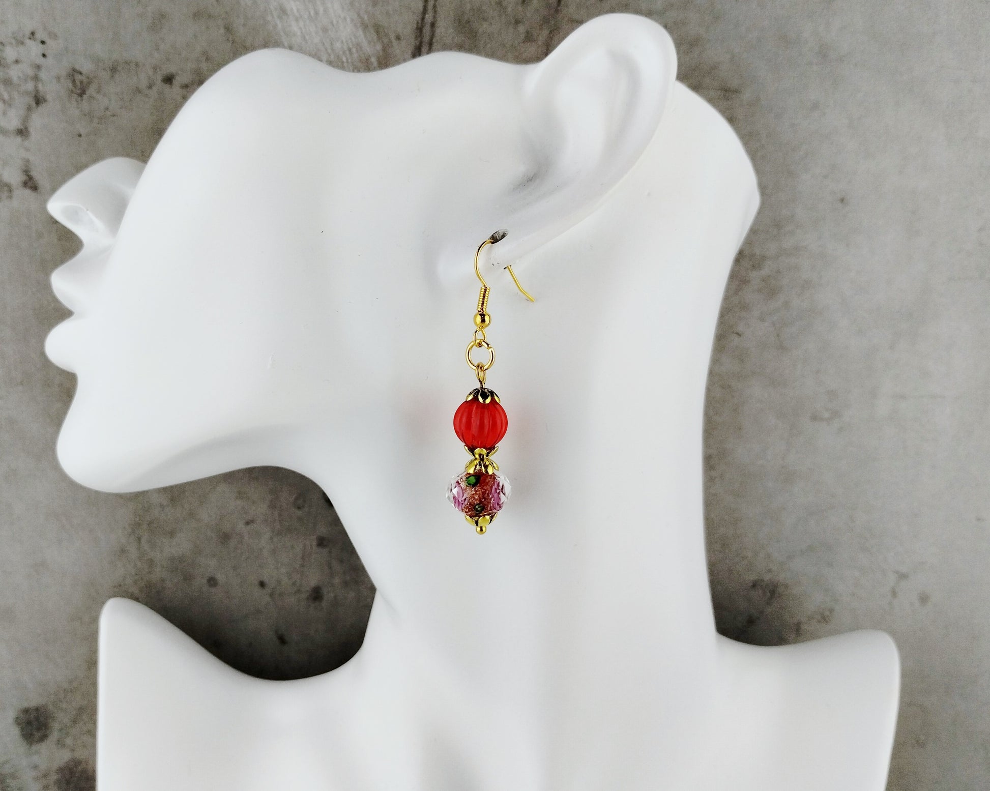 A single dangling earring displayed on a white mannequin head against a gray textured background. The earring has a gold fishhook wire, a round red bead, a small dark spacer, and a faceted pink crystal charm.