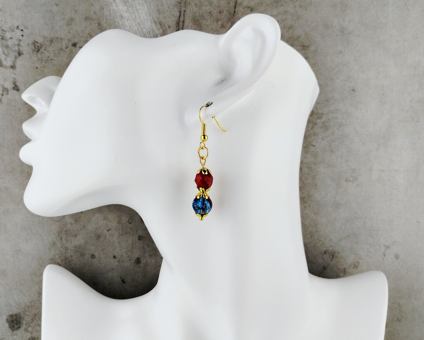 A single dangling earring displayed on a white mannequin head against a gray textured background. The earring has a gold-tone hook, a round dark red bead, and a faceted blue bead with gold detailing.