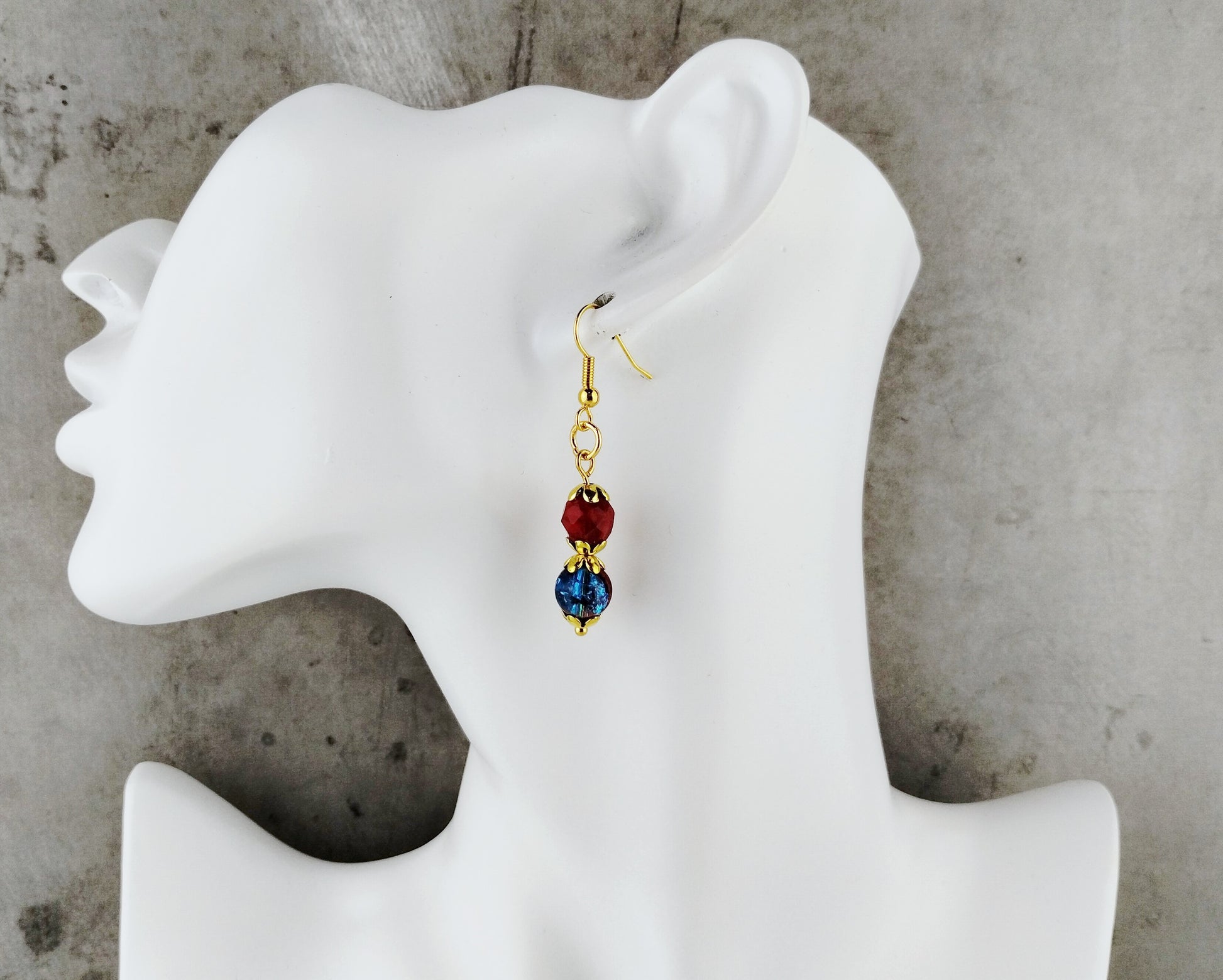 A single dangling earring displayed on a white mannequin head against a gray textured background. The earring has a gold-tone hook, a round dark red bead, and a faceted blue bead with gold detailing.