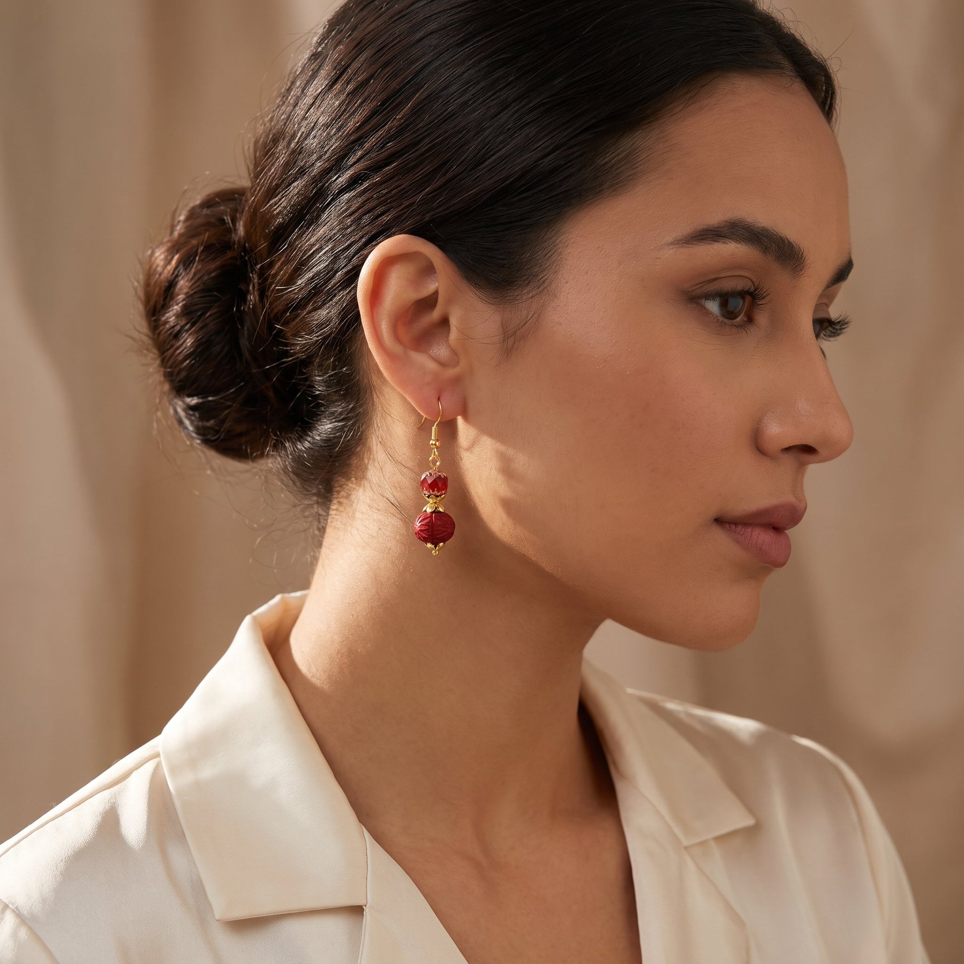 A woman in profile wearing a pair of gold drop earrings with red beads, set against a soft beige background.