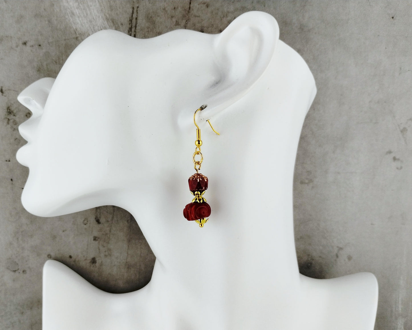 A single dangling earring displayed on a white mannequin head against a gray concrete-like background. The earring has gold-tone hardware and two dark red beads.