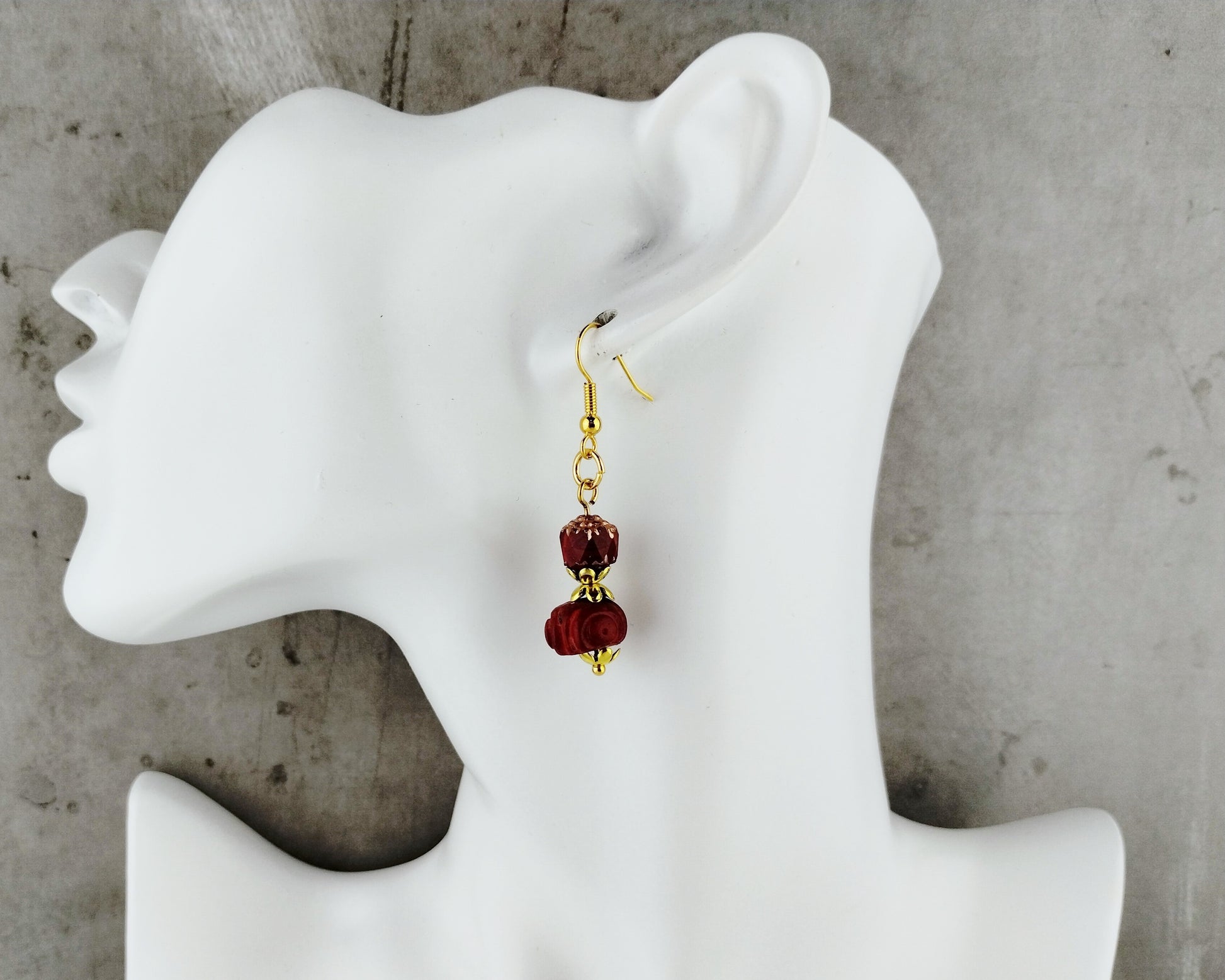 A single dangling earring displayed on a white mannequin head against a gray concrete-like background. The earring has gold-tone hardware and two dark red beads.