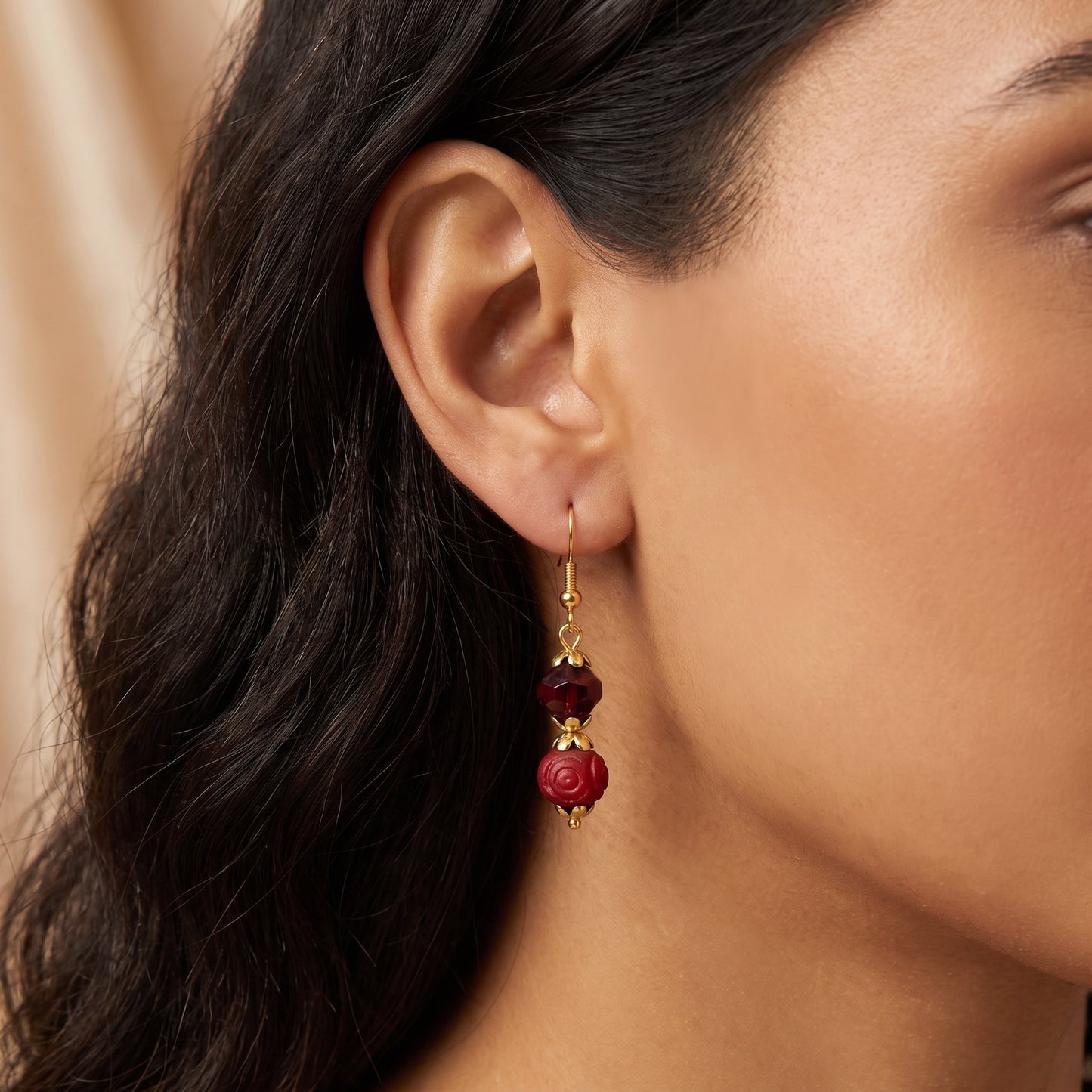 A close-up of a woman's ear wearing a dangling earring with gold-toned metal and deep red faceted beads.