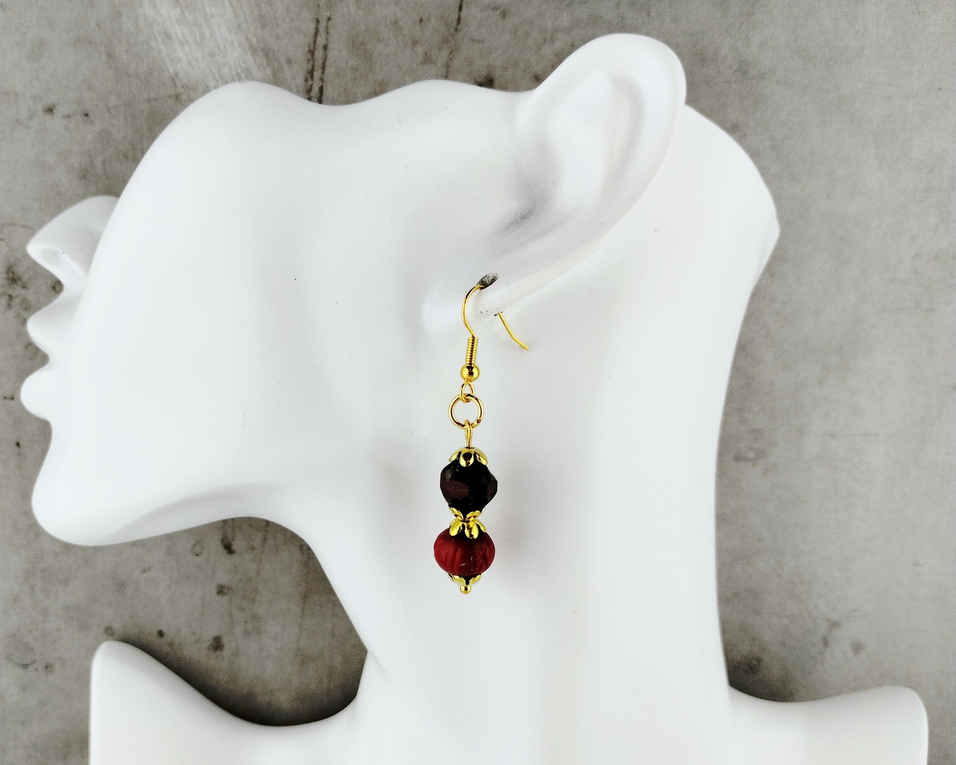 A single dangling earring displayed on a white mannequin head against a gray concrete-like background. The earring has a gold-tone fishhook wire, a small black bead, and a larger red bead with gold-tone caps.