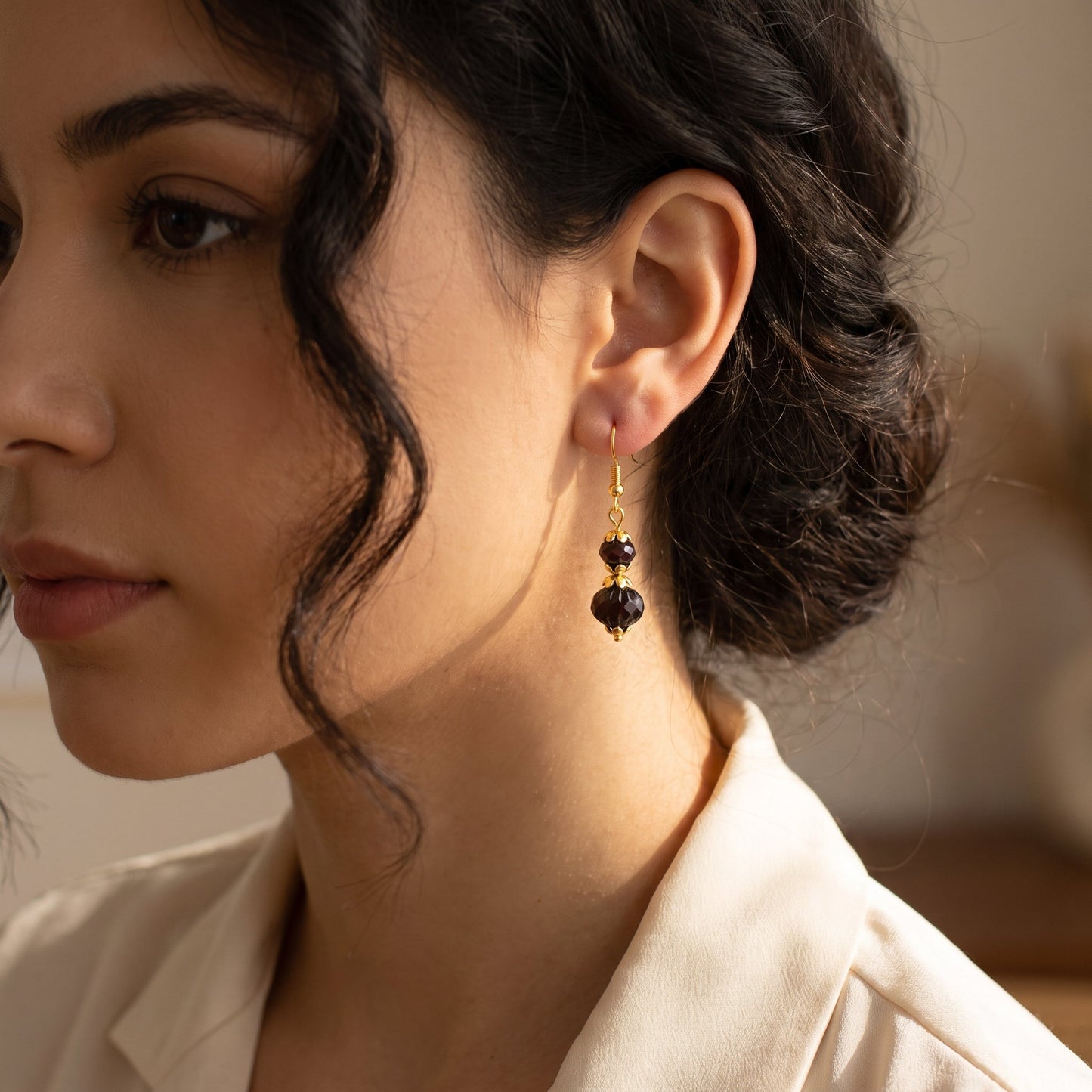A close-up of a woman wearing a dangling earring with two dark faceted beads and gold-toned metal details, paired with a cream-colored blouse.