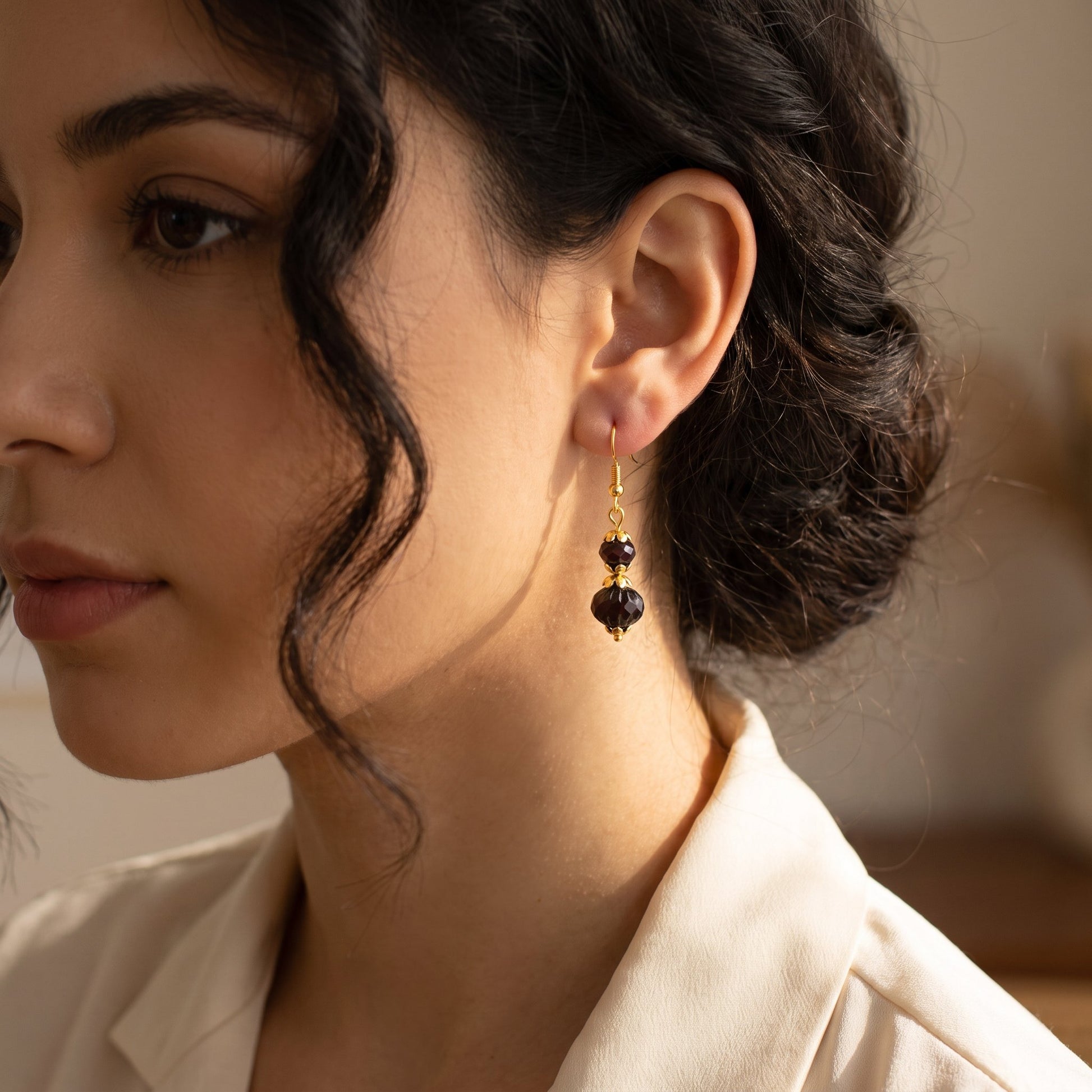 A close-up of a woman wearing a dangling earring with two dark faceted beads and gold-toned metal details, paired with a cream-colored blouse.