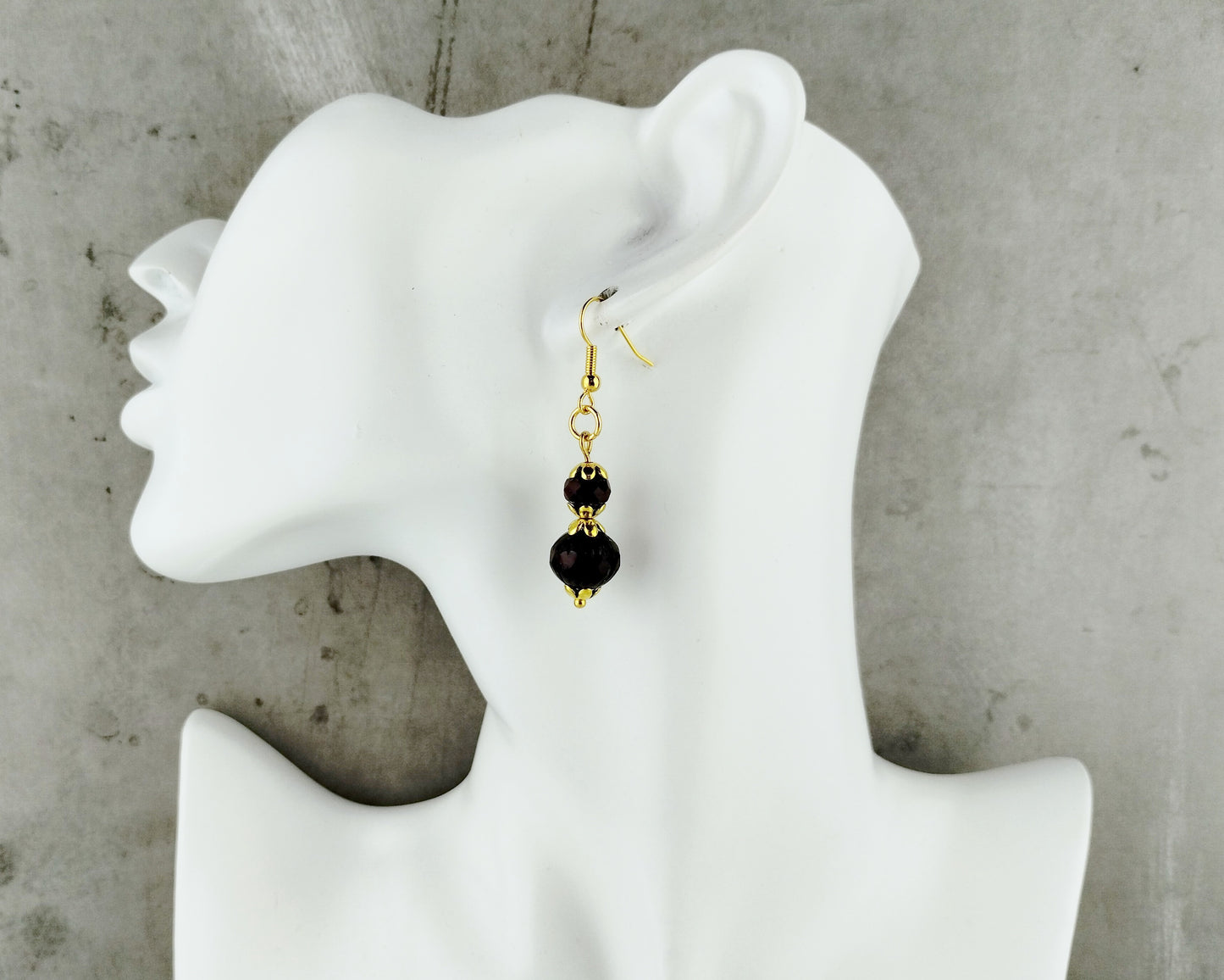 A single drop earring displayed on a white mannequin head against a gray concrete-textured background. The earring has a gold fishhook clasp with two small gold spacer beads and a larger faceted red bead dangling at the bottom.