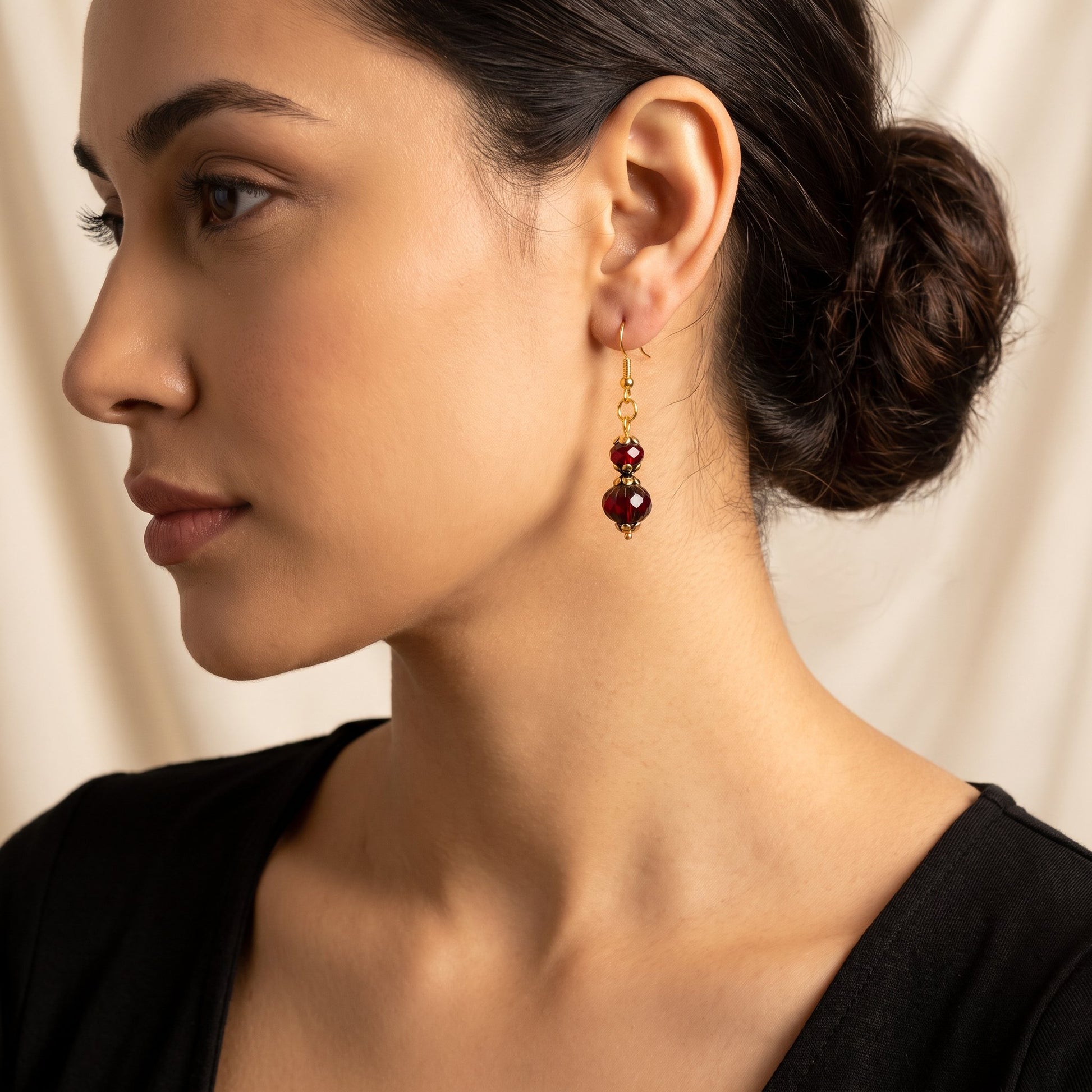A close-up side profile of a woman wearing a dangling gold earring with two dark red beads, set against a soft beige fabric background.