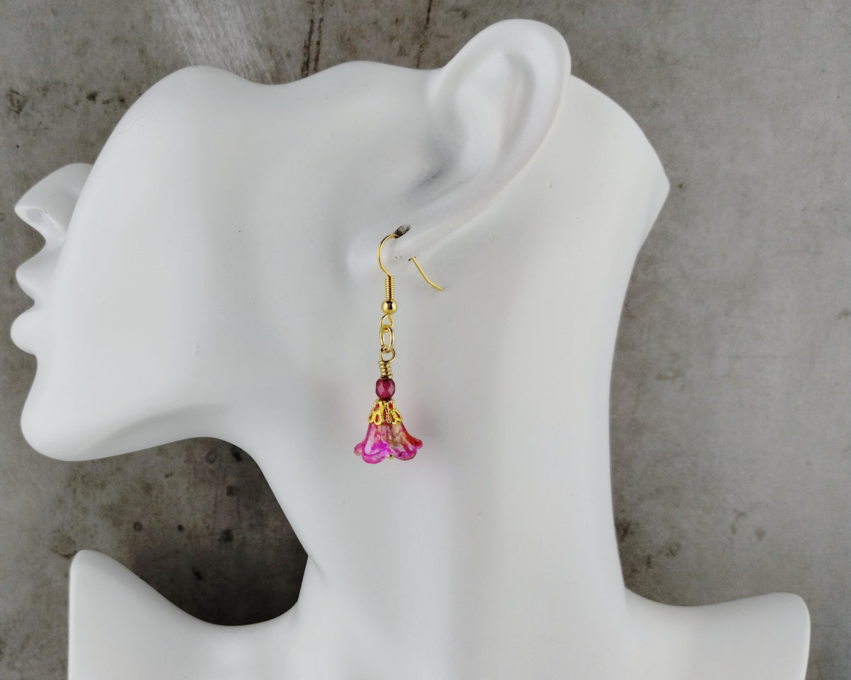 A single dangling earring displayed on a white mannequin head against a gray concrete-textured background. The earring has a gold fishhook wire, a small dark red bead, and a pink flower-shaped charm with gold detailing.