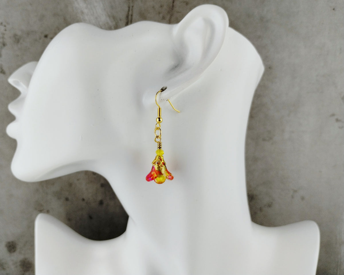 A single dangling earring displayed on a white mannequin head against a textured gray background. The earring has a gold-toned fishhook wire and a small floral drop with yellow and red translucent petals.