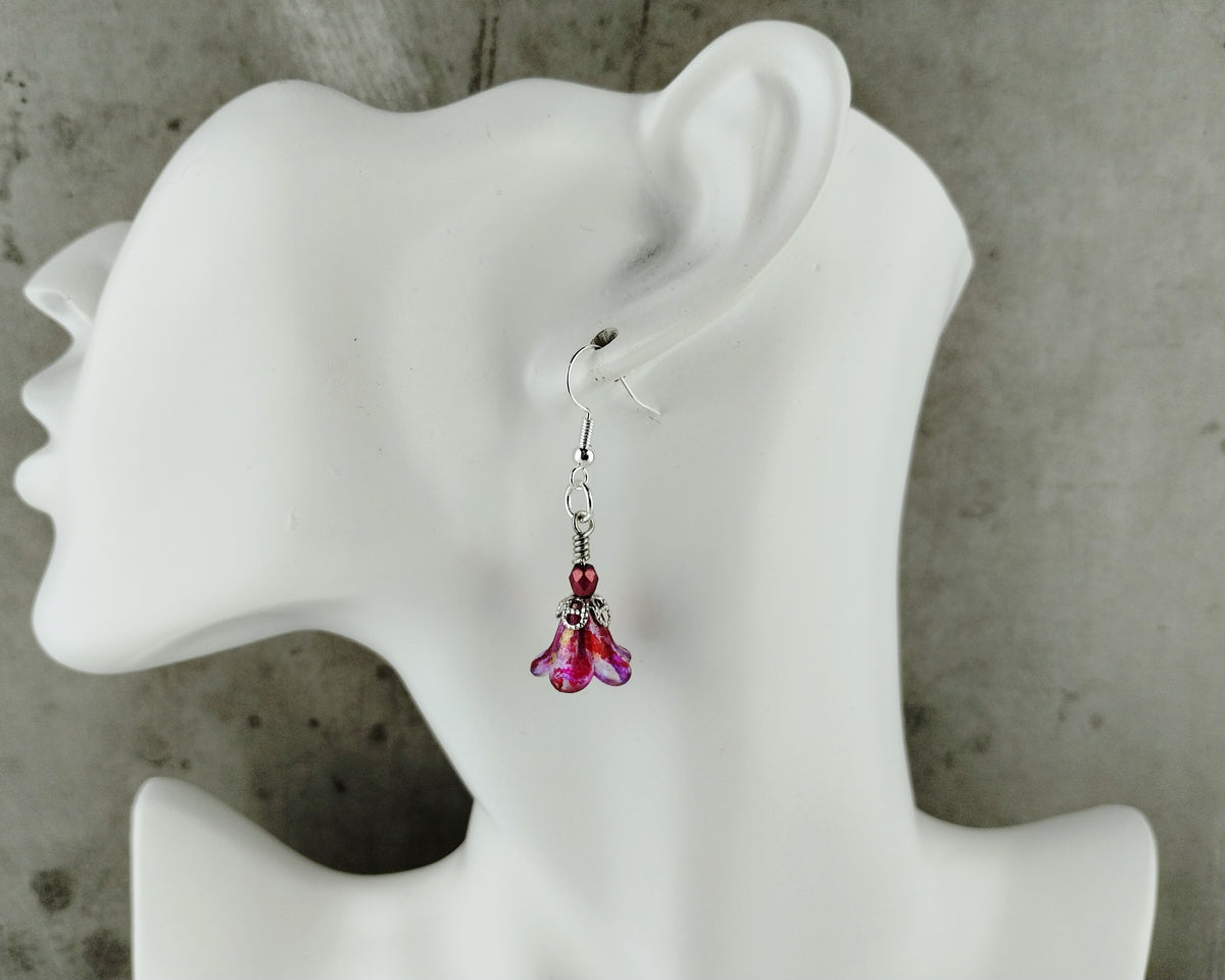 A single dangling earring displayed on a white mannequin head against a textured gray background. The earring has a silver-toned fishhook  dangling flower-like drops in pink and purple.
