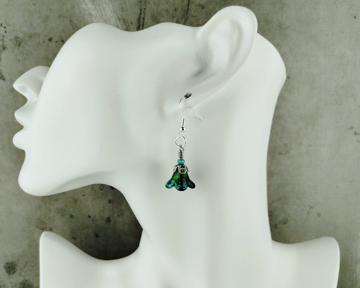 A single dangling earring displayed on a white mannequin head against a gray concrete-textured background. The earring has a silver-toned fishhook back with a small silver bead and a dangling bell-shaped charm in iridescent green and blue tones.