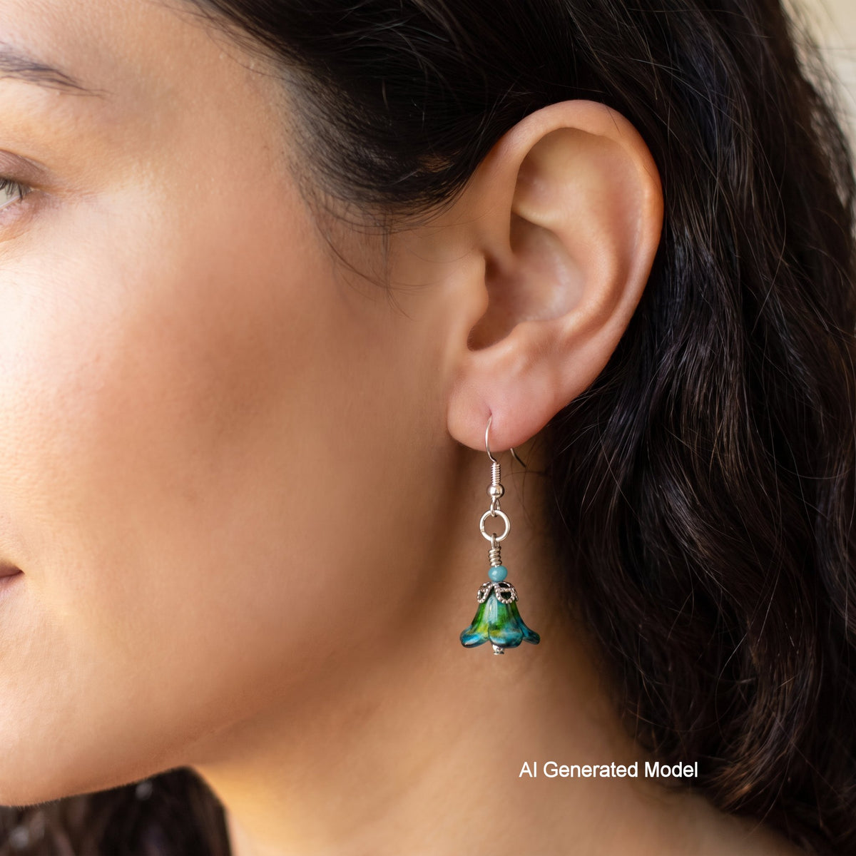 A close-up side profile of a person wearing a dangling earring. The earring has a silver-toned fishhook and ring, with a small green and blue enamel bell-shaped charm.