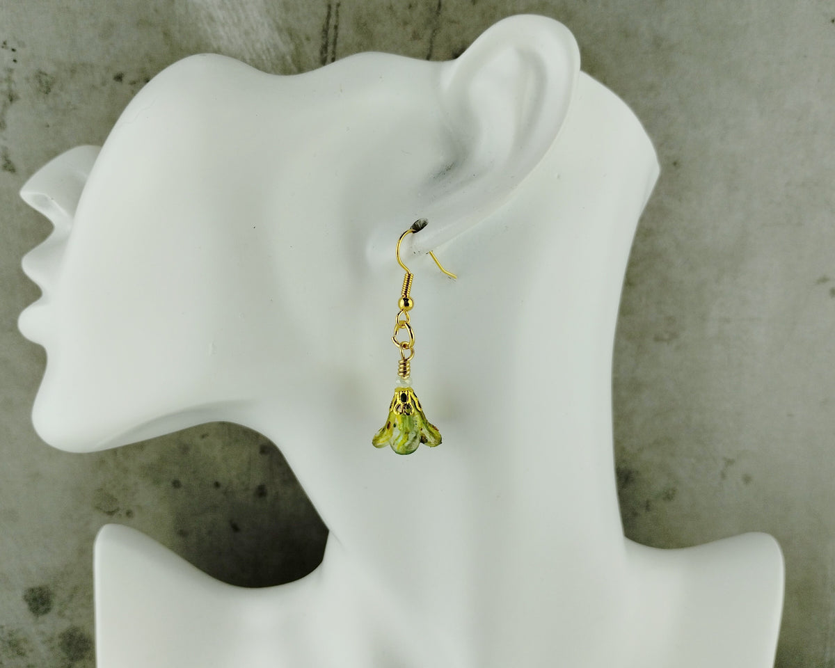 A single gold dangle earring with a small green bell-shaped charm, displayed on a white female mannequin head against a textured gray concrete background.