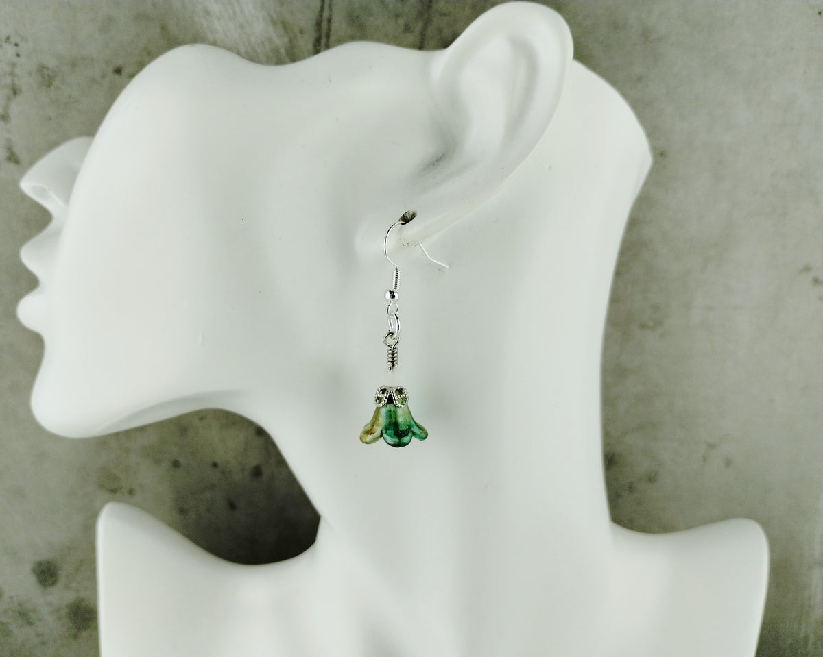 A single dangling earring displayed on a white mannequin head against a textured gray background. The earring has a silver-toned fishhook back and a small dangling charm shaped like a green frog with yellow-green accents.