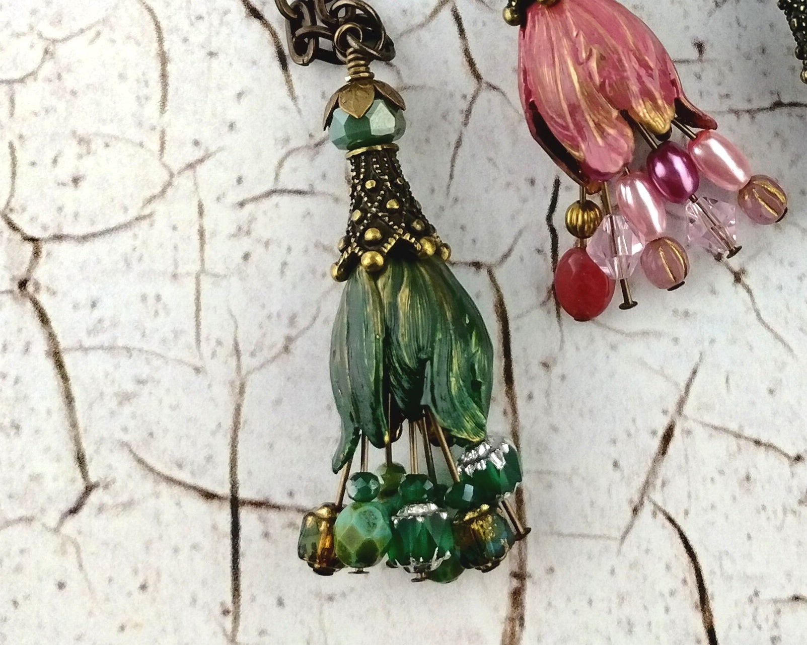 A close-up of a handmade tassel pendant necklace featuring green glass beads shaped like leaves, accented with bronze metal details and small green and silver beads, set against a textured off-white background.