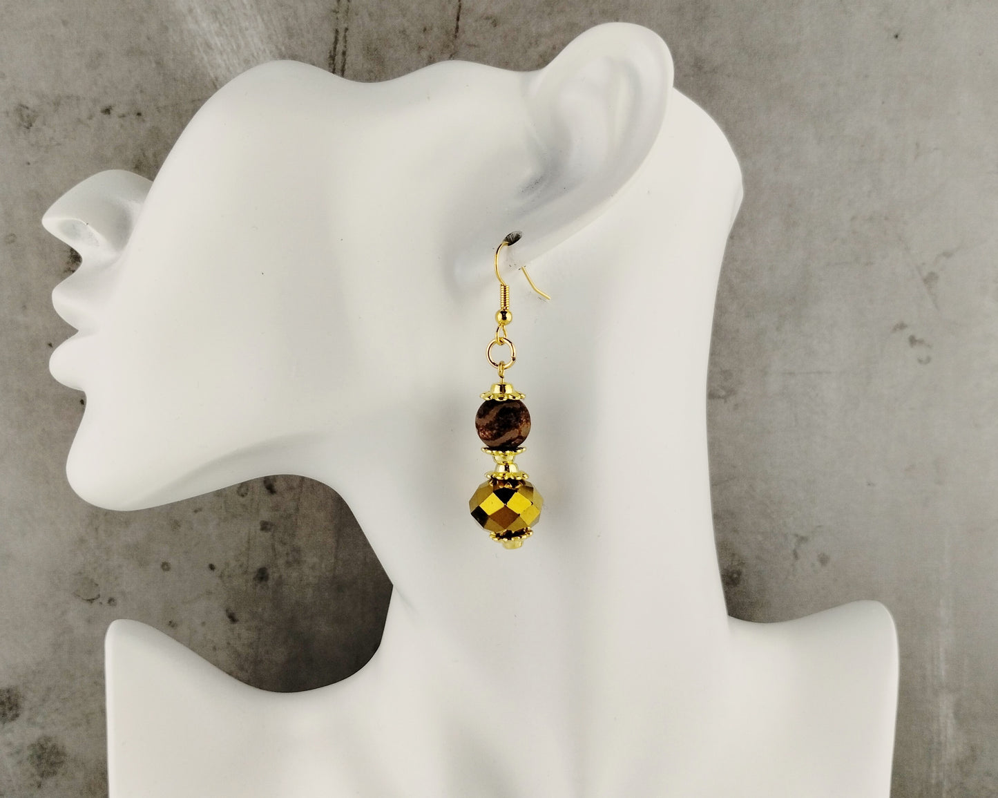 Gold earrings with brown  beads on a mannequin ear against a gray background