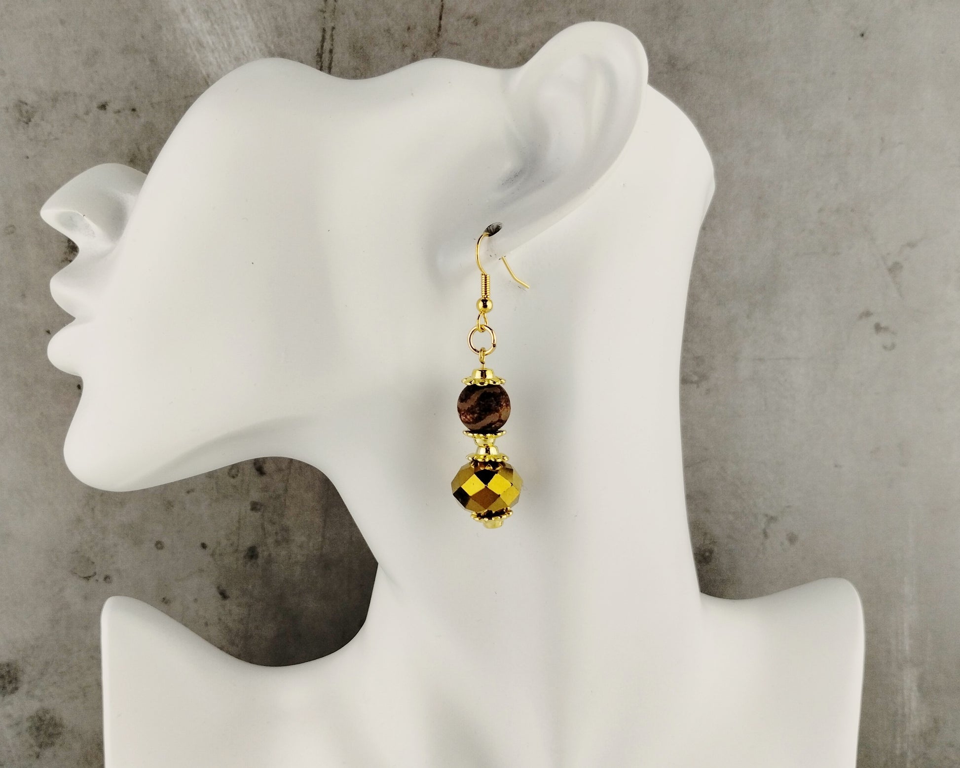 Gold earrings with brown  beads on a mannequin ear against a gray background