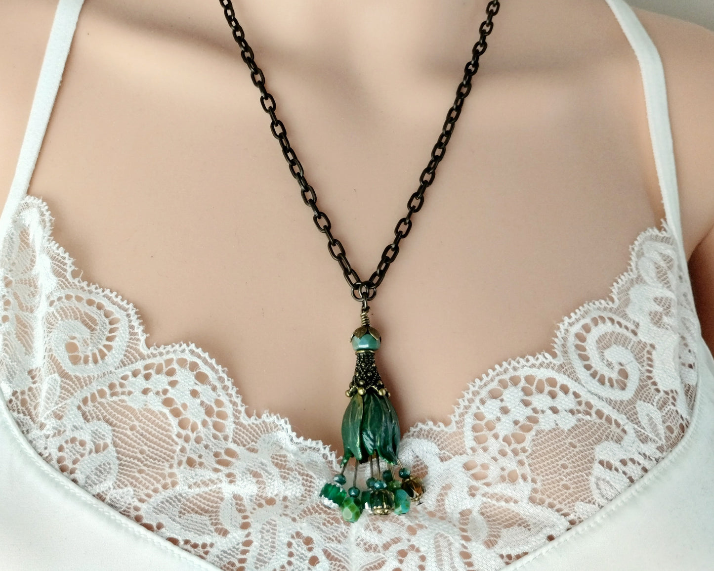 A close-up of a necklace worn on a mannequin or model torso, featuring a brass chain and a green beaded pendant with a tassel-like drop, styled over a white lace bra.