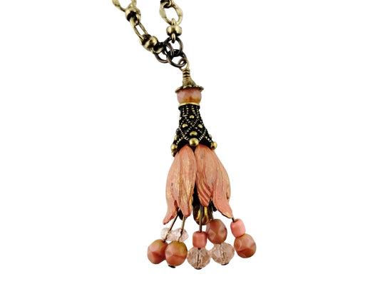 A bohemian-style pendant necklace featuring a cluster of peach-pink flower petals wrapped in black and gold beaded wire, hanging from an antique bronze chain.