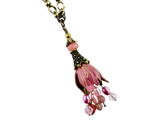 A close-up of a dangling necklace pendant with a pink flower-shaped charm, pink beads, and a gold-toned chain on a plain white background.